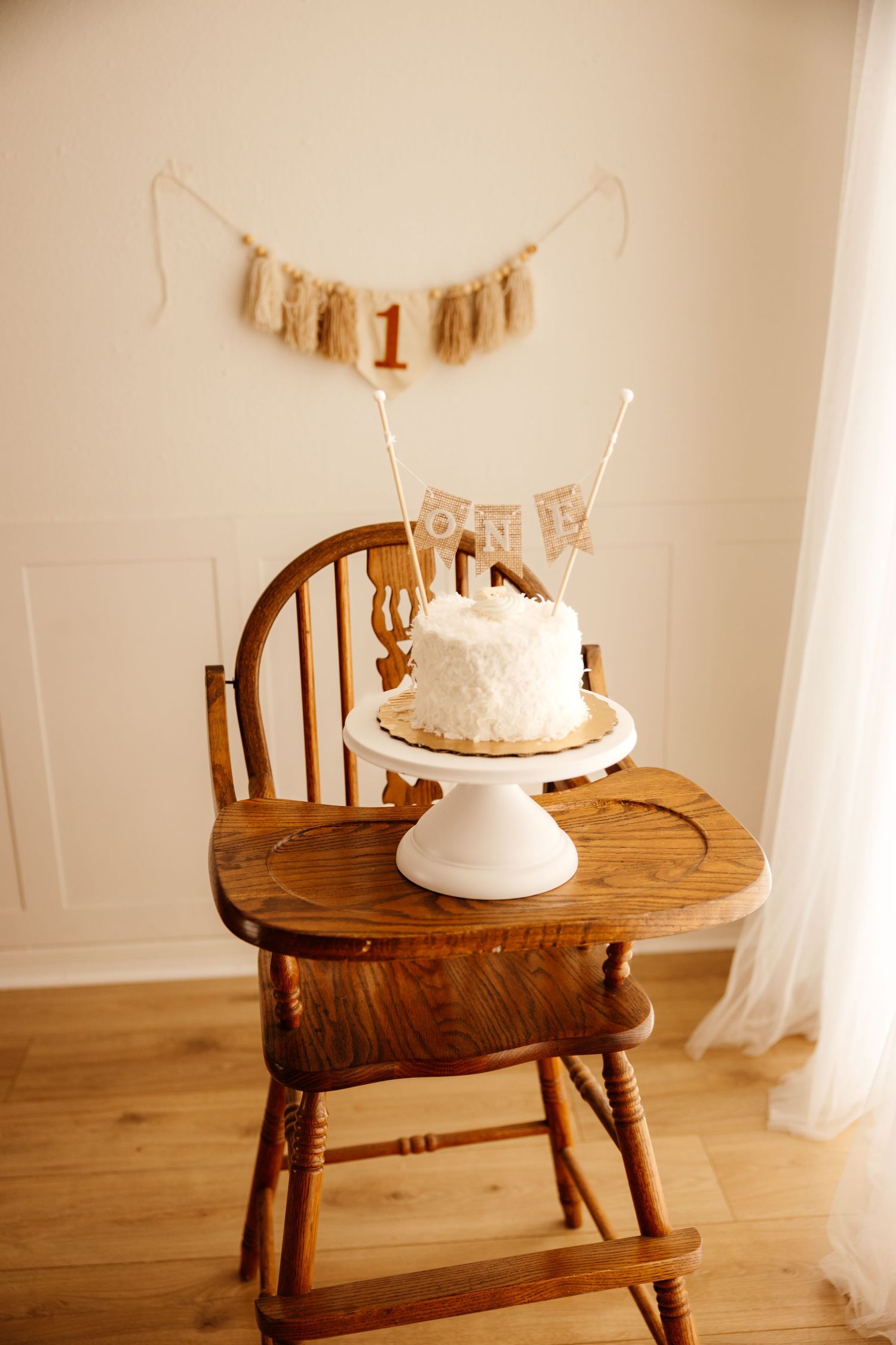 White frosted first birthday cake on a wooden high chair with ONE topper and number 1 bunting banner decoration.