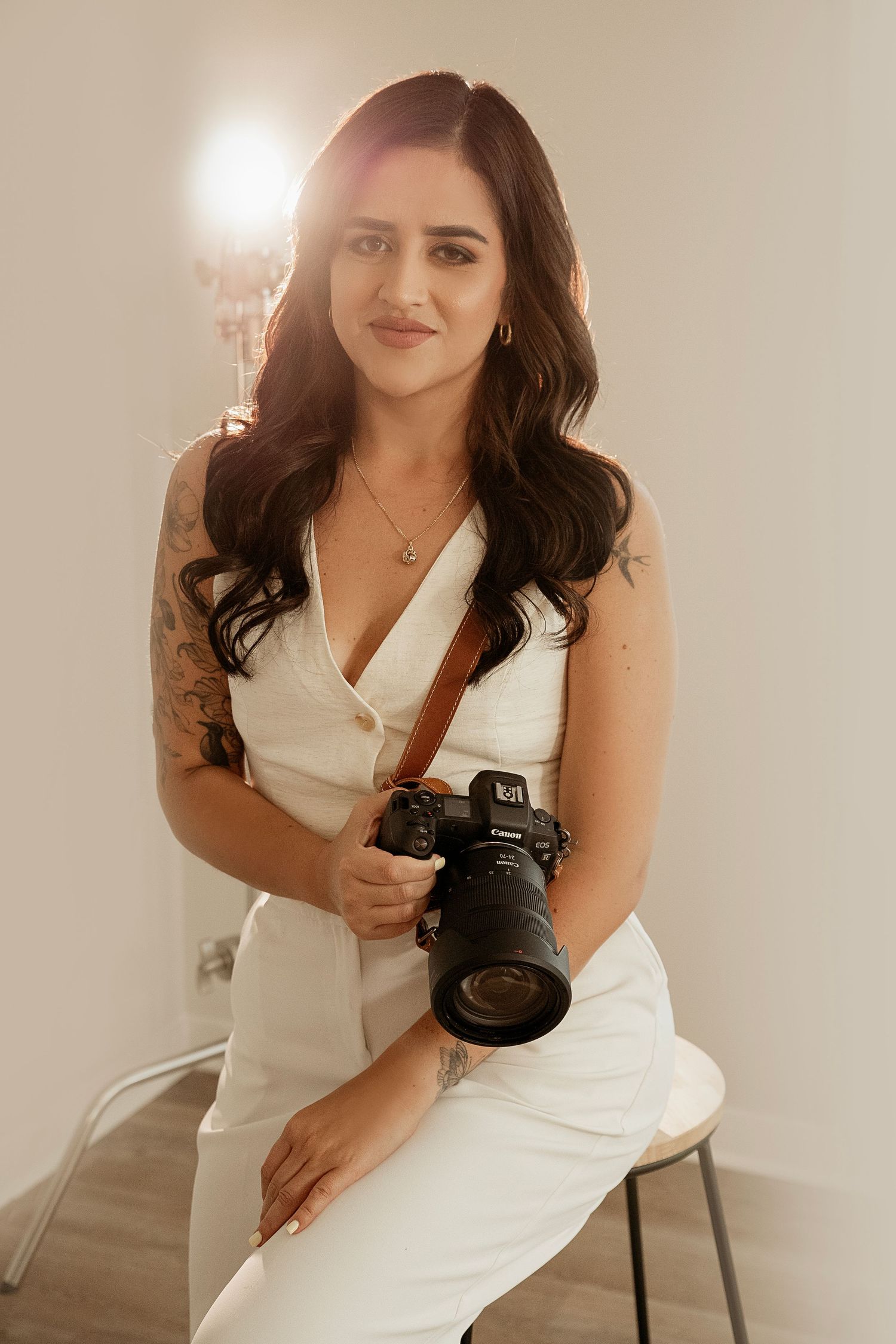 Professional photographer in a white dress holds a professional DSLR camera with a large lens while posing on a chair.