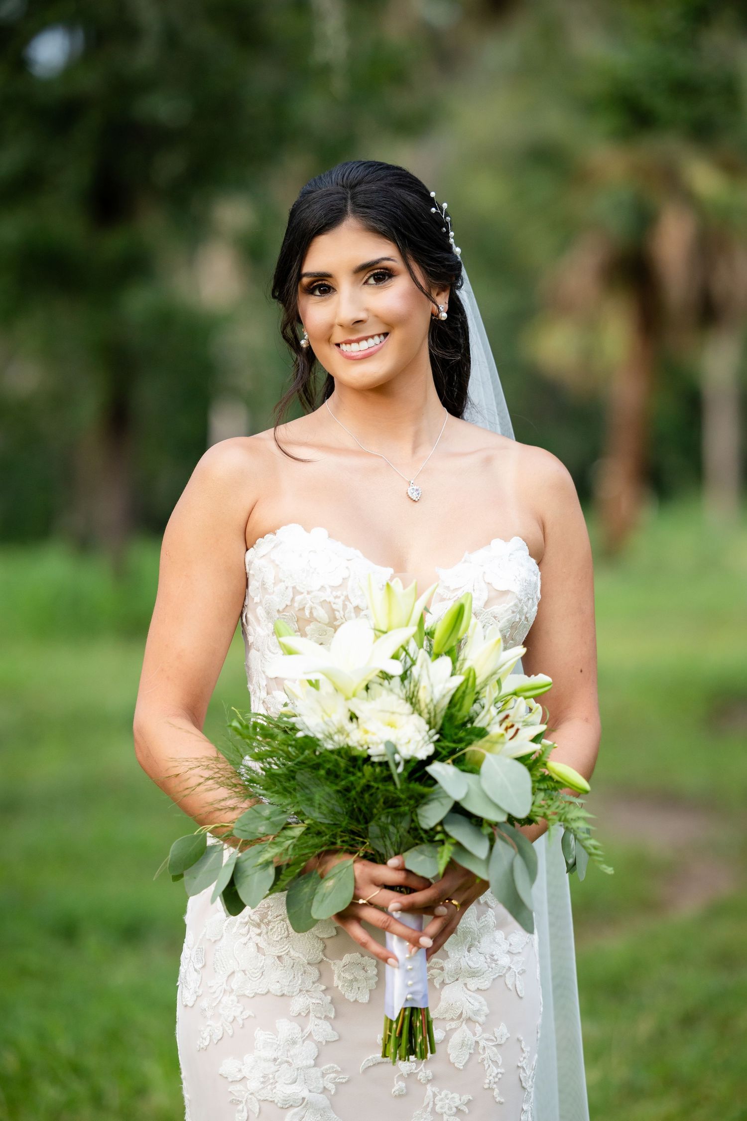 Wedding portrait showing bride in white lace gown holding elegant bouquet of white lilies and greenery in outdoor setting.