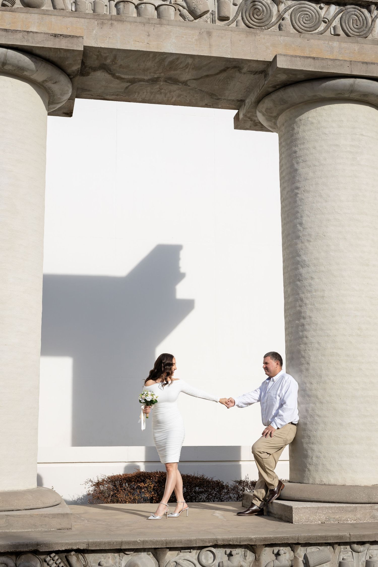 Couple sitting on concrete ledge at ancient Greek-style columns with dramatic shadows and light.