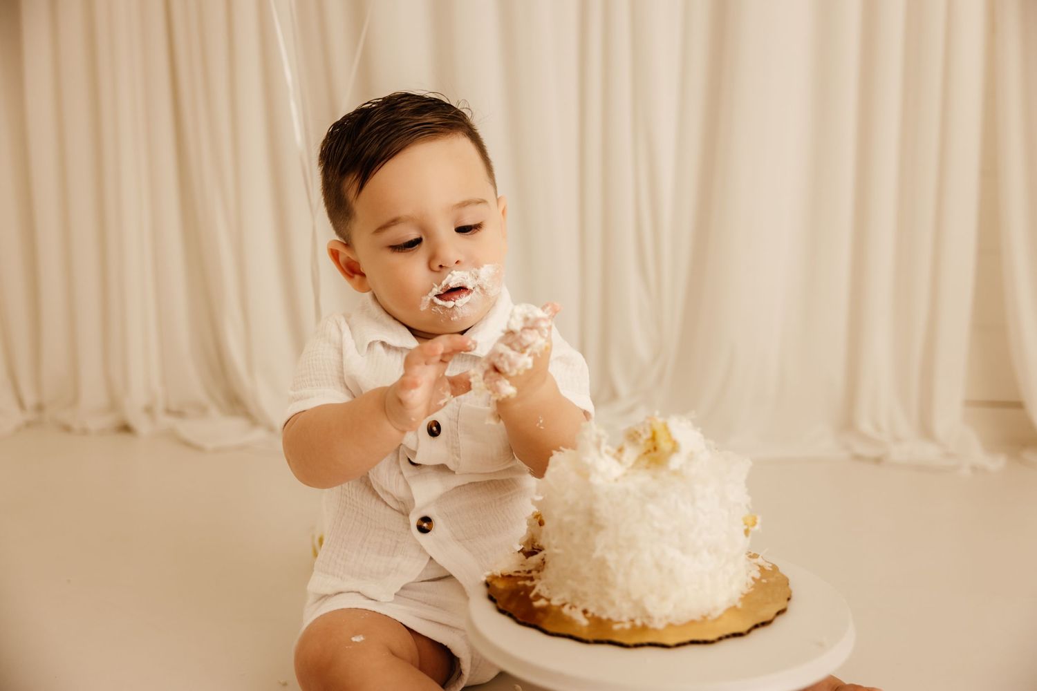 A baby boy in a white romper smashes his first birthday cake, covered in white frosting, against a neutral backdrop.