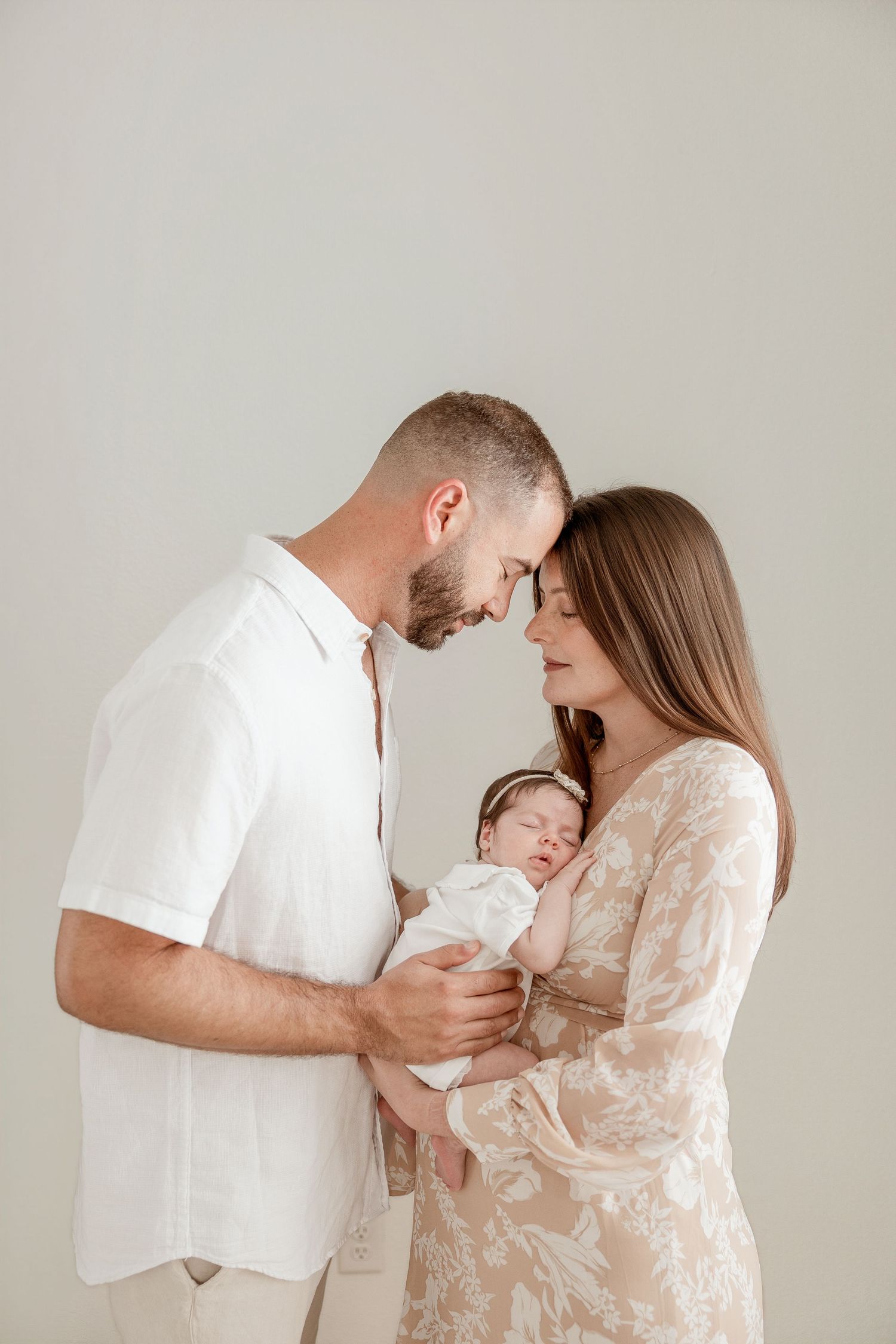 A couple tenderly holds their sleeping newborn baby, touching foreheads in an intimate studio portrait.