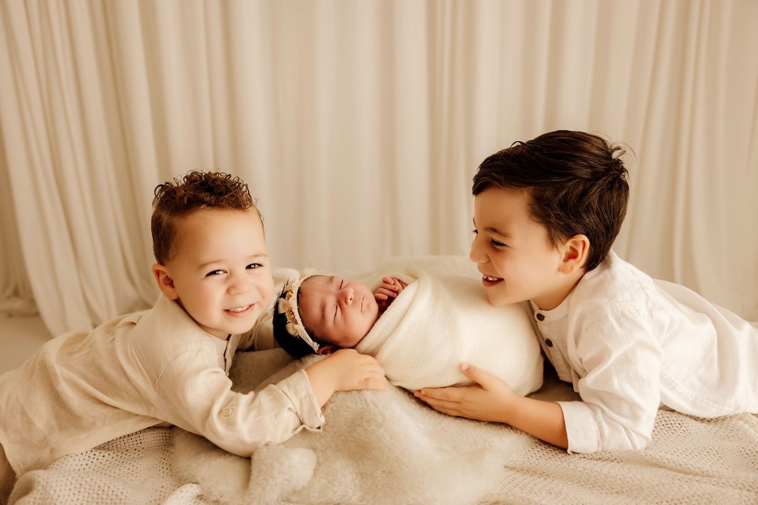 Two young brothers smile lovingly at their newborn sibling wrapped in white on a soft blanket.