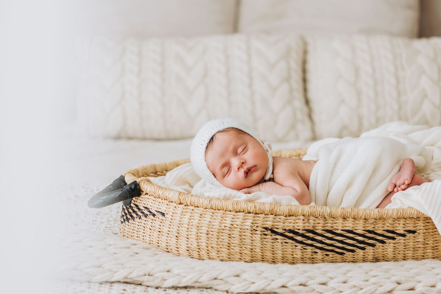 Newborn baby sleeping peacefully on white bedding next to a woven basket in natural light.