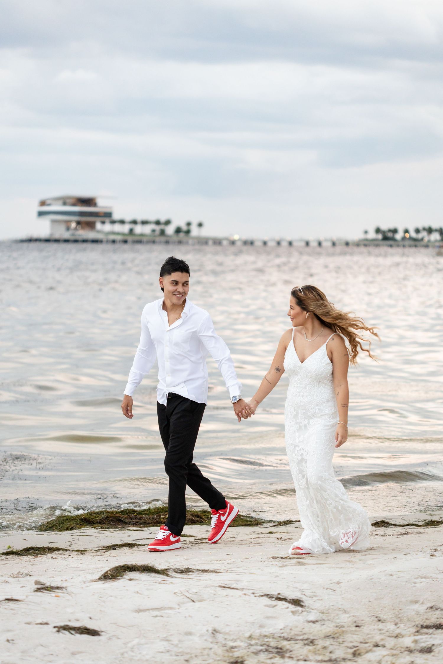 A couple runs playfully on a beach at sunset wearing white and dark attire with red sneakers.