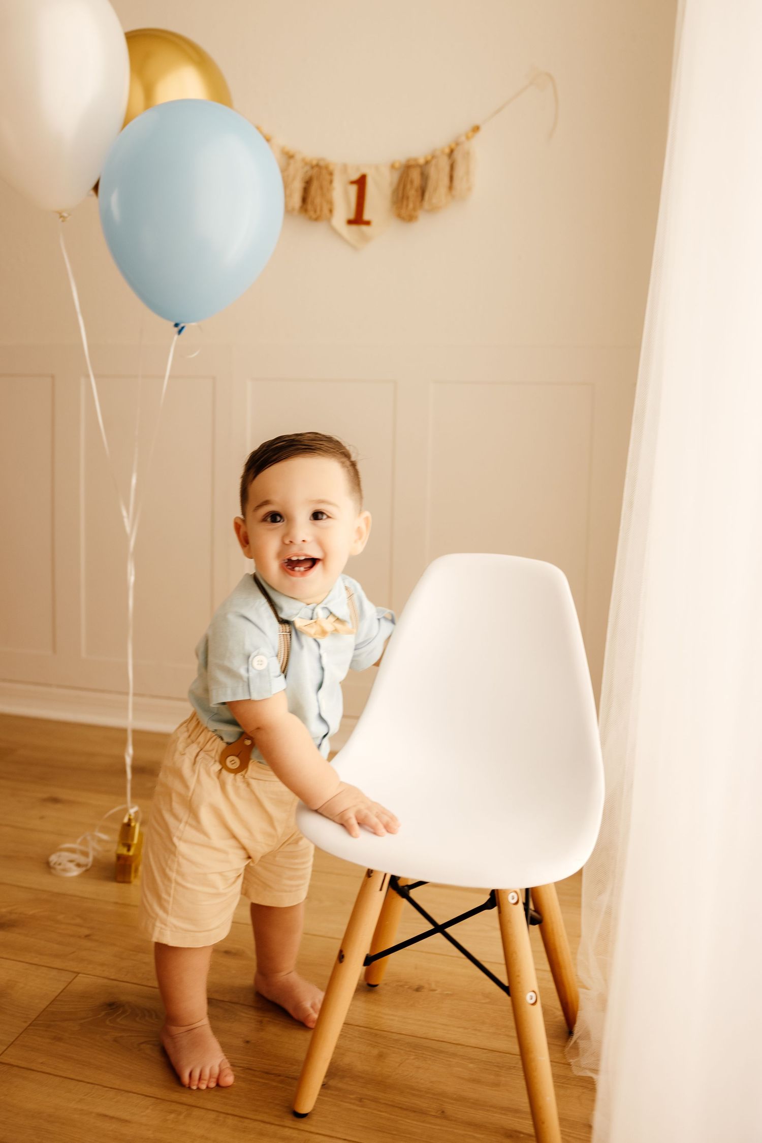 Smiling baby boy celebrates first birthday with balloons and number one banner, standing by white chair on wood floor.