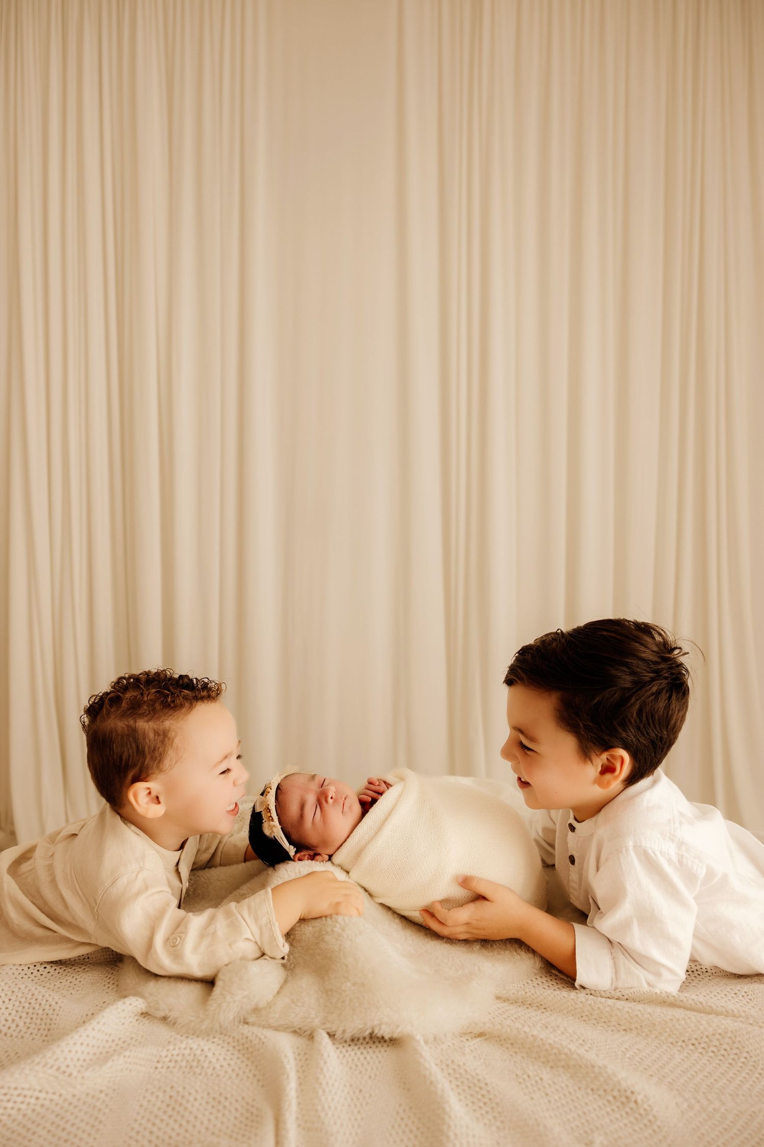 Two young brothers in white outfits lovingly gaze at their swaddled newborn sibling on a cream blanket.