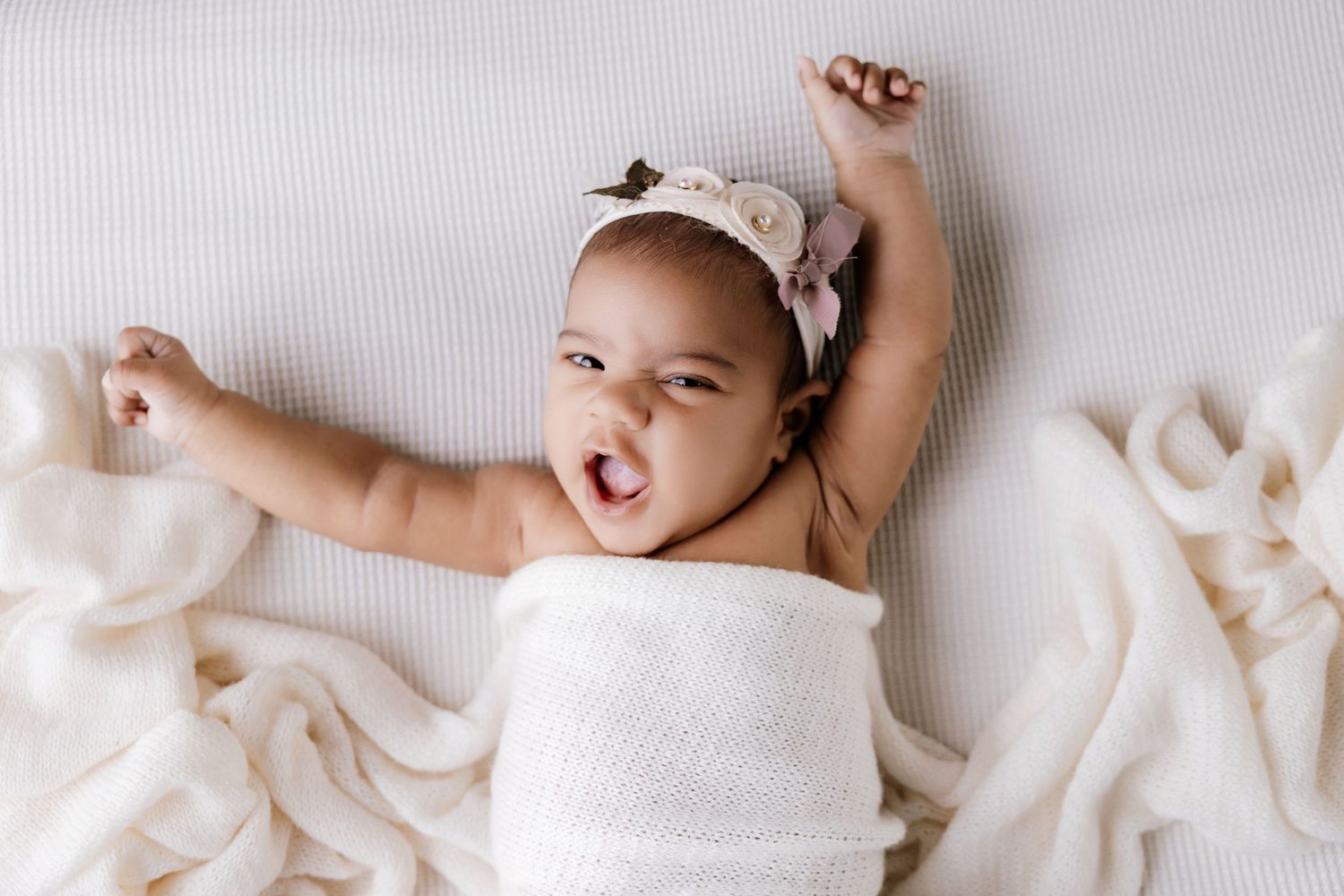 Adorable baby girl in white wrap and floral headband stretching and smiling while lying on a soft blanket.