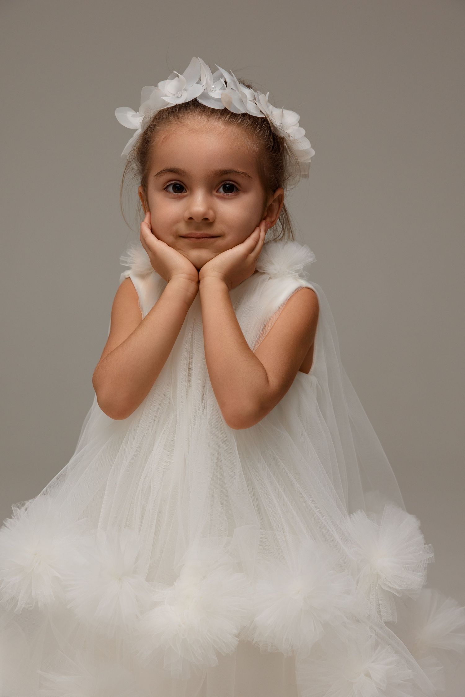 Young girl in white tulle dress and floral headpiece poses with hands on cheeks against gray background.