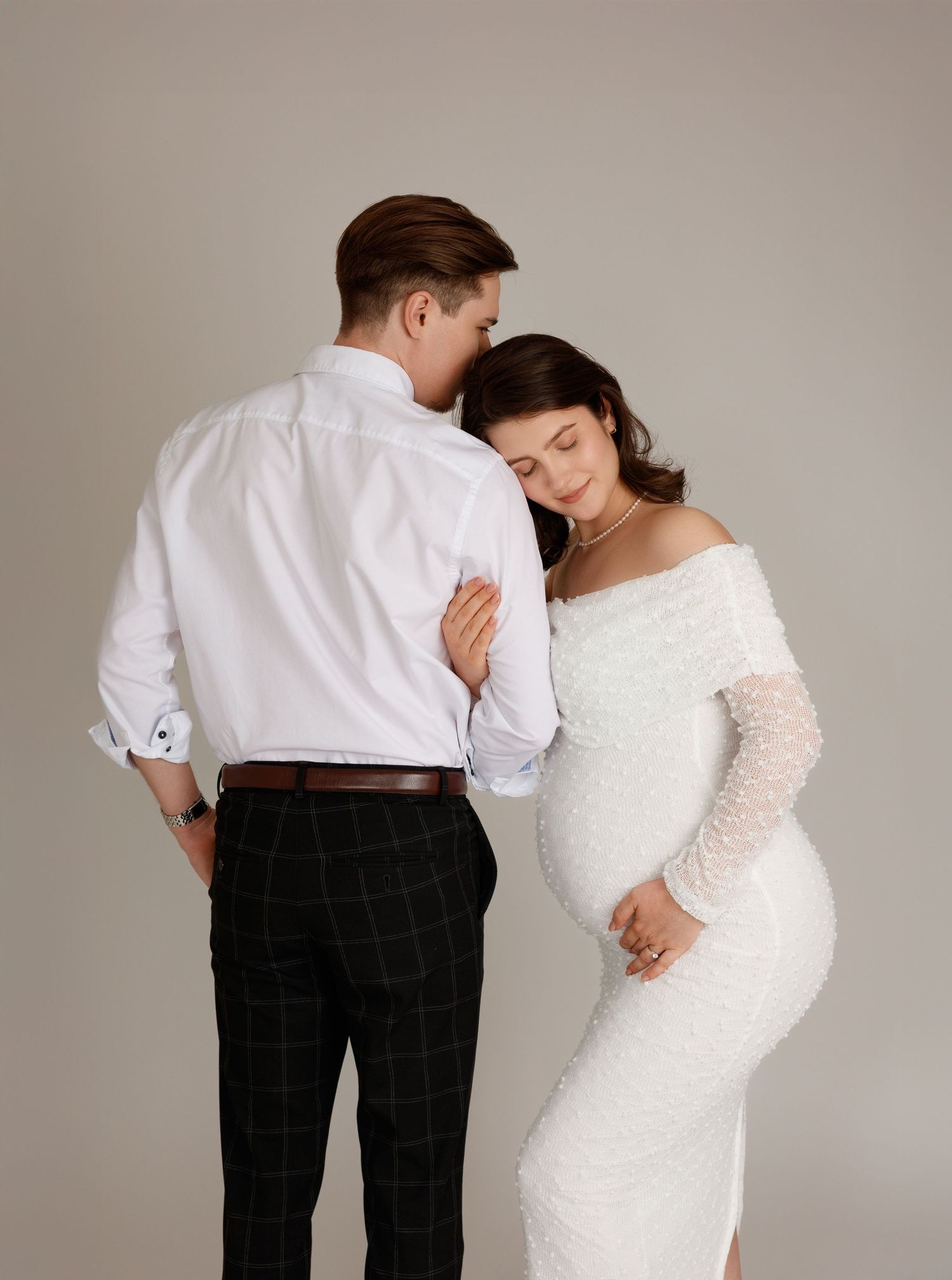 Elegant maternity photo of a couple in white and plaid, sharing a tender moment in a studio setting.