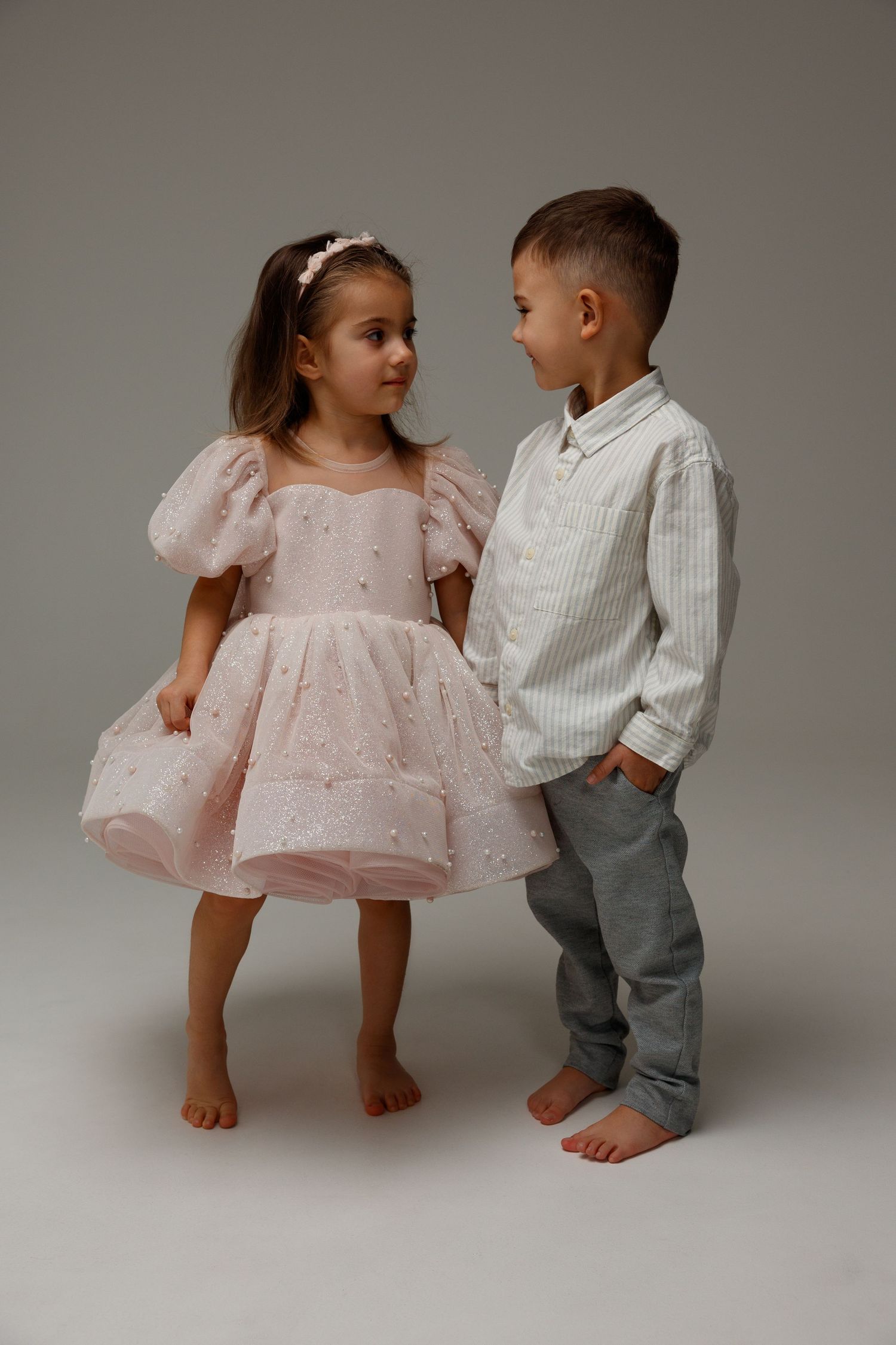 Two young children dressed up, a girl in a pink tulle dress and a boy in a striped shirt, standing barefoot facing each other.