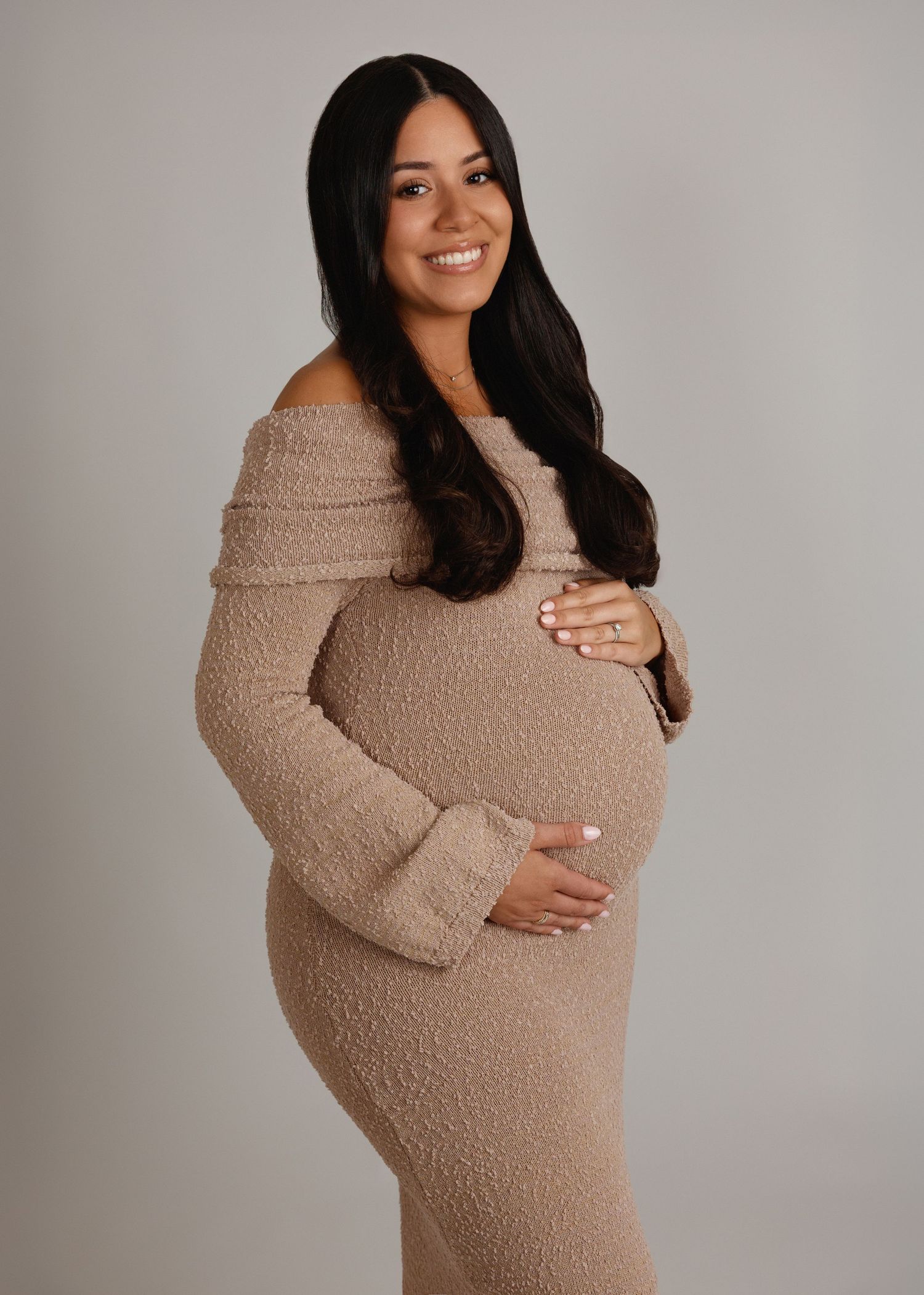 Smiling pregnant woman in beige off-shoulder textured dress cradling baby bump, dark hair, studio portrait.