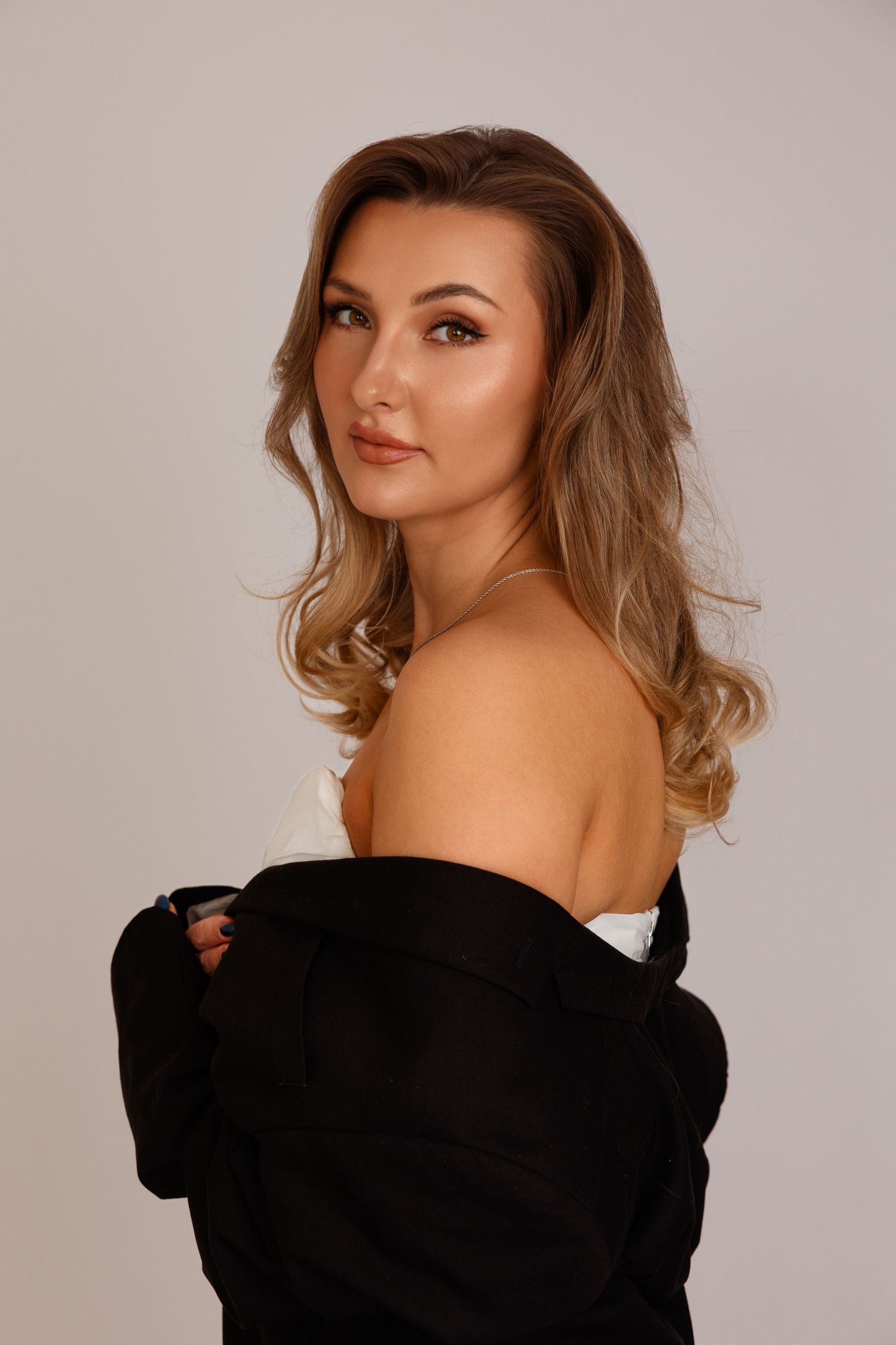 Young woman with blonde wavy hair posing in black jacket with bare shoulders against gray background.