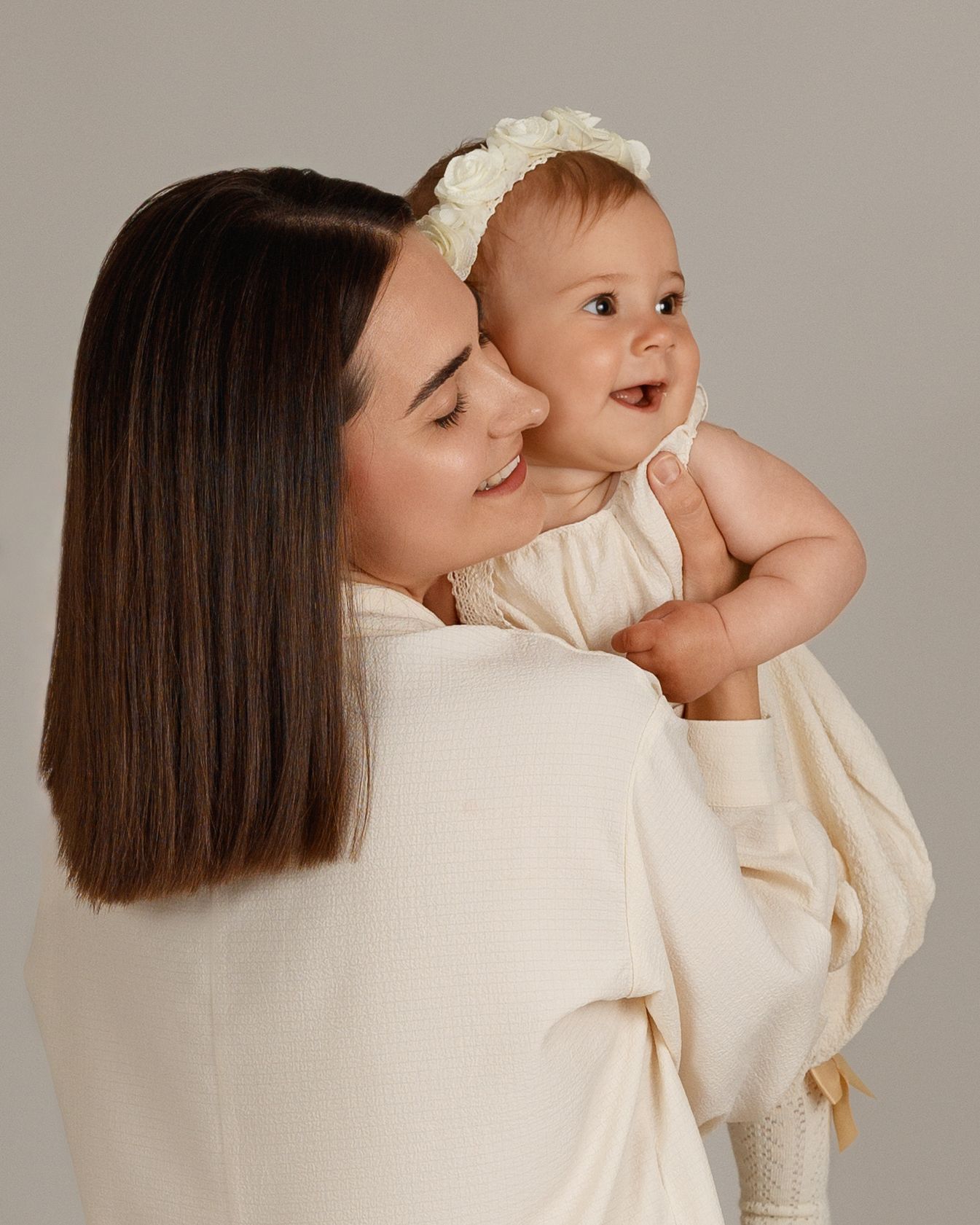 A mother in cream attire holds her smiling baby girl wearing a floral headband against a neutral background.