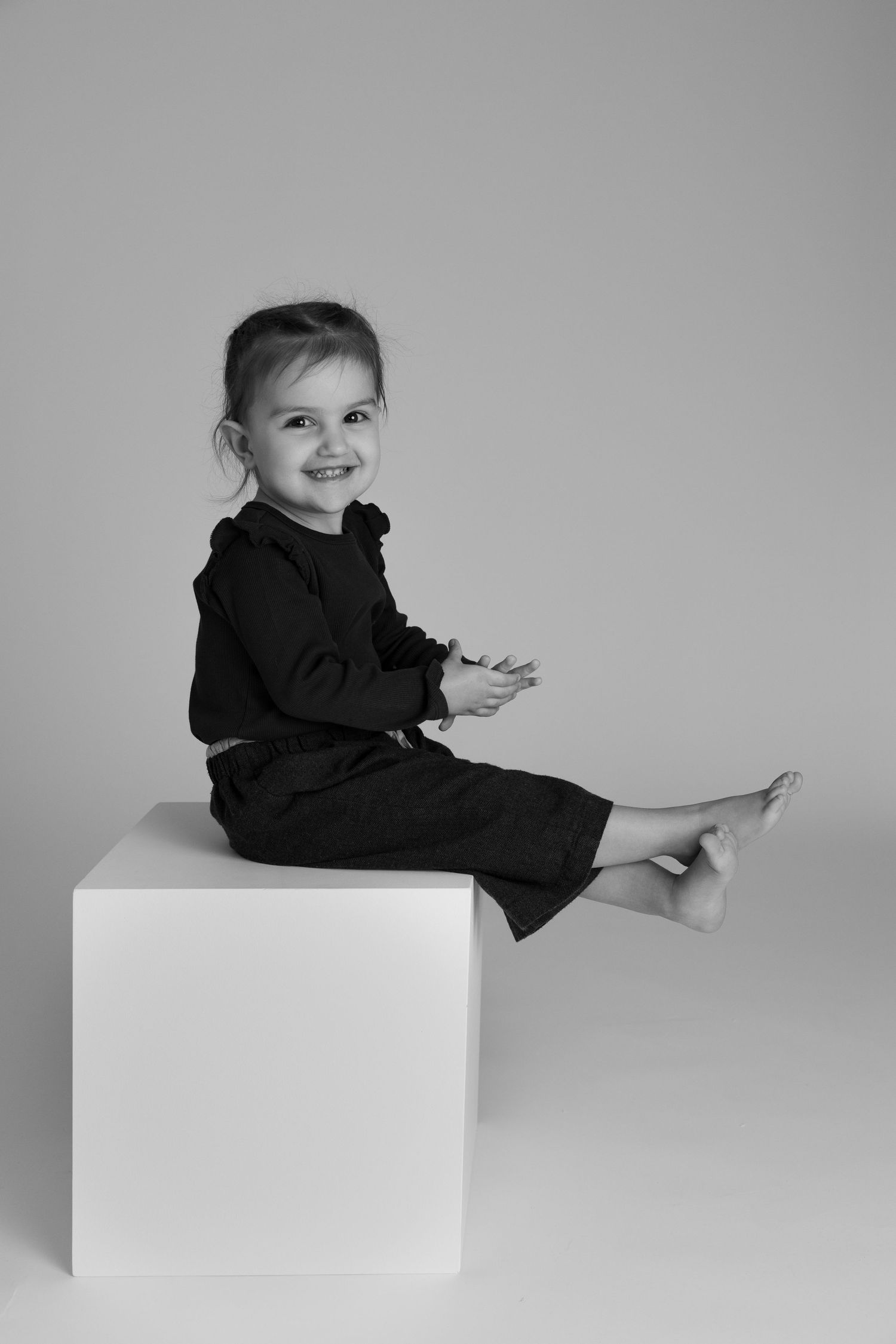 Black and white portrait of a smiling young girl in dark outfit sitting barefoot on a white cube.