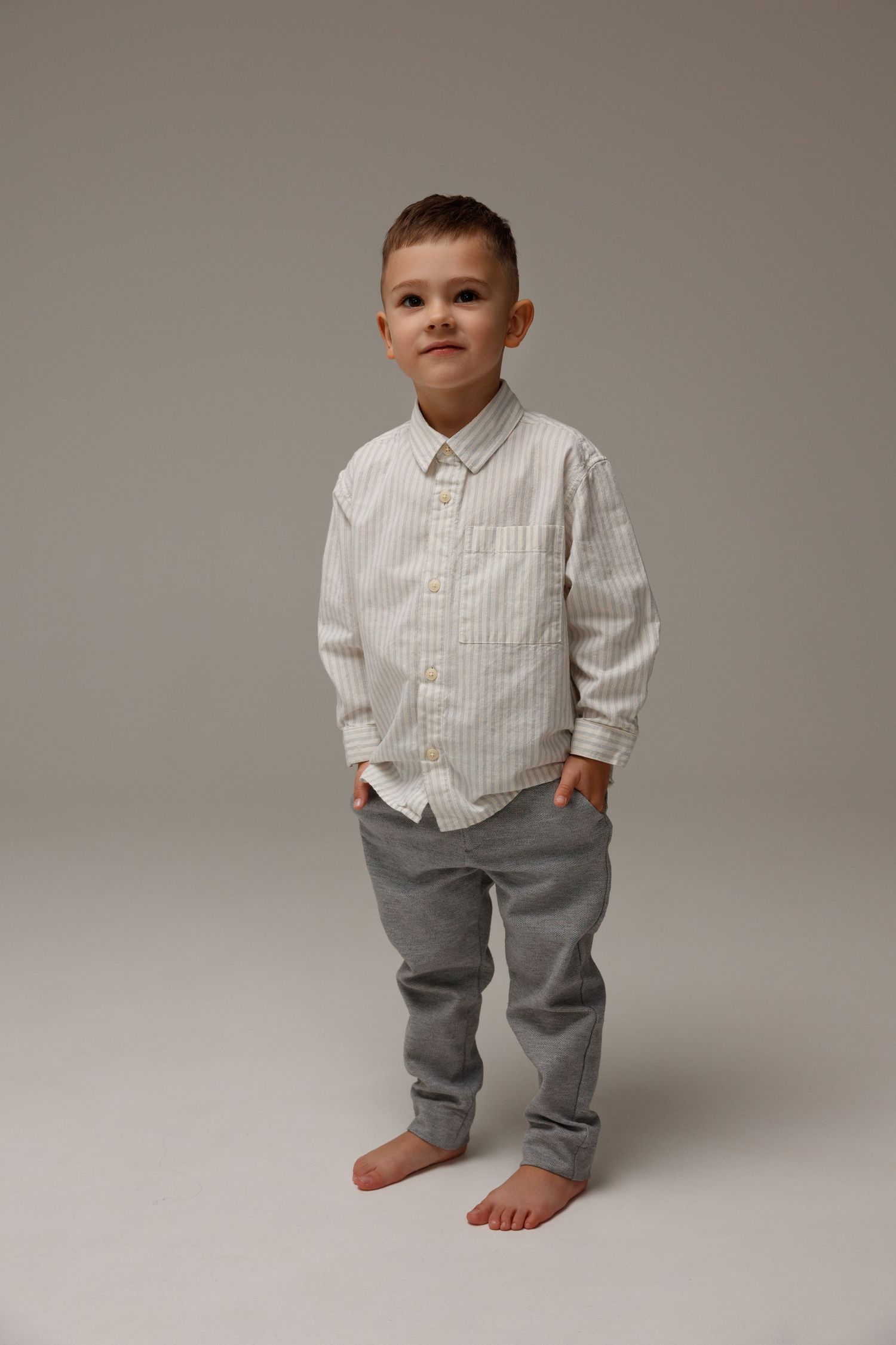 Young boy in striped shirt and gray pants standing barefoot against a light gray background.