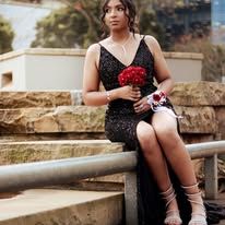 Young woman in black sequin dress holding red roses bouquet, seated on steps wearing silver heels for prom.