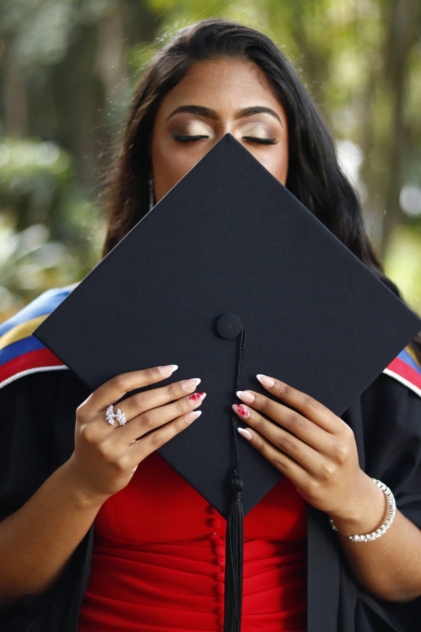 Young woman in graduation gown holding black mortarboard cap over her face with eyes closed.