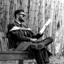 Graduate in cap and gown reads document on bench surrounded by bamboo in black and white photo.