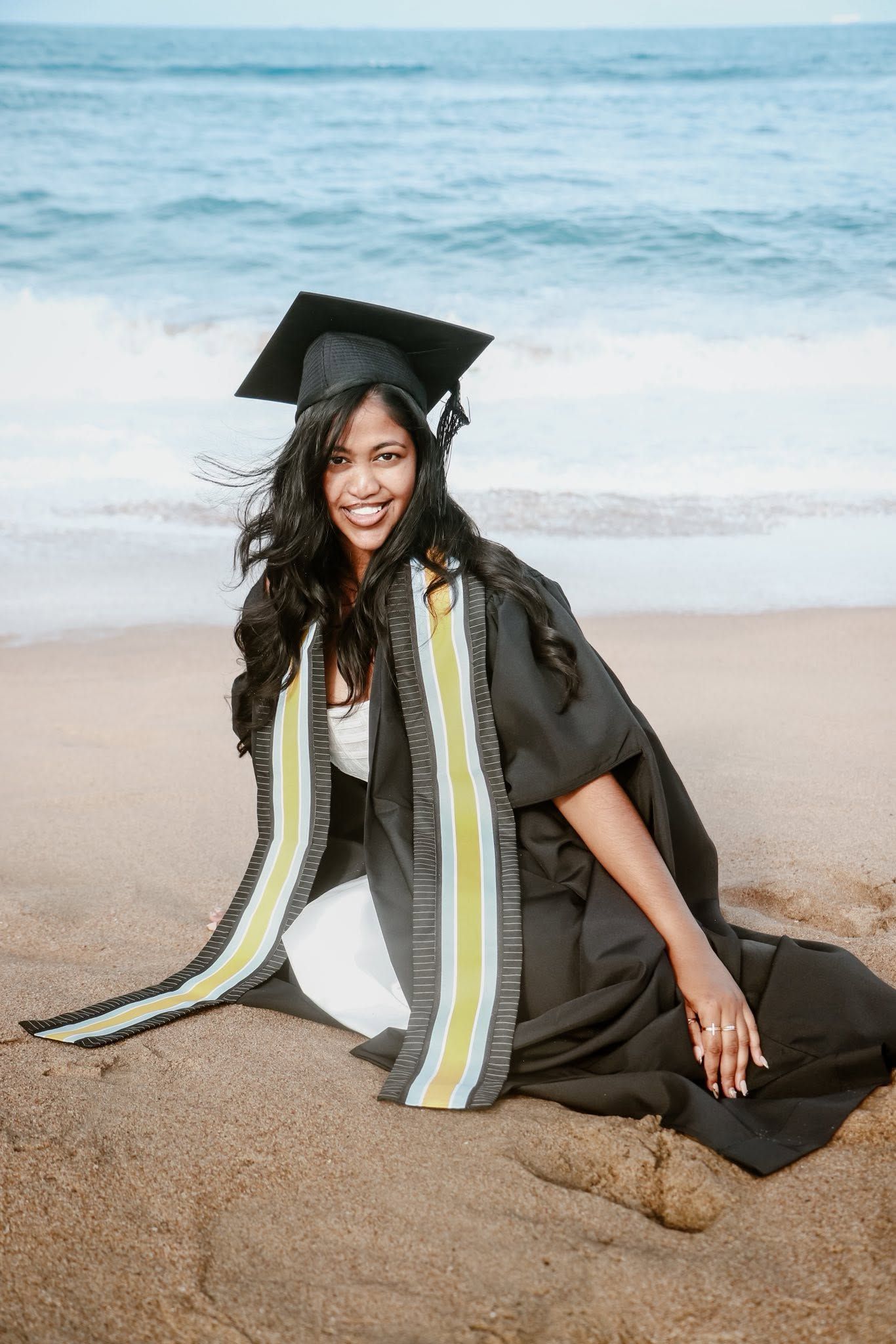 Smiling graduate in black cap and gown with yellow sash sitting on sandy beach with ocean waves behind her.