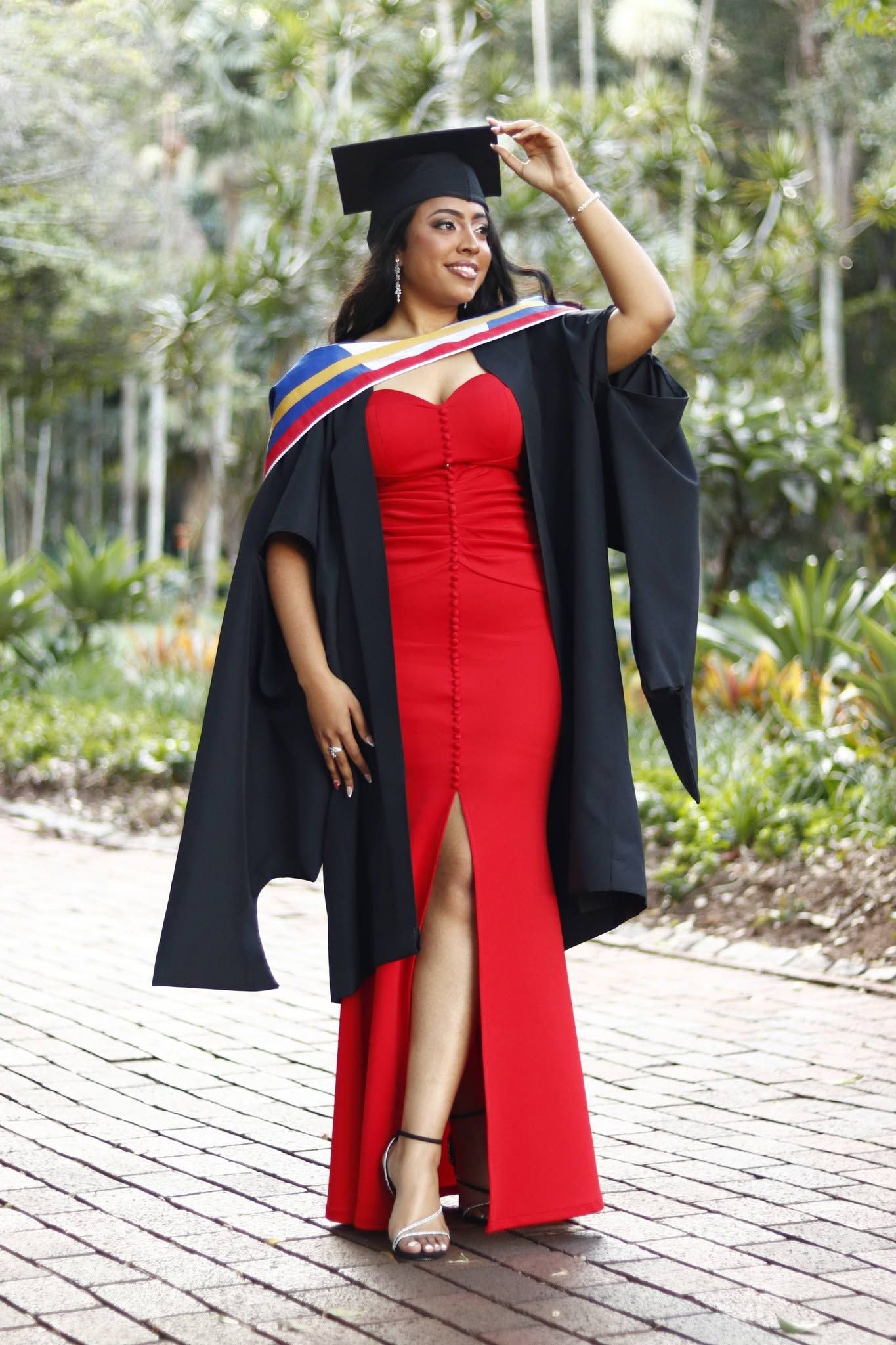 A smiling graduate in red dress and black gown holds her mortarboard cap outdoors.