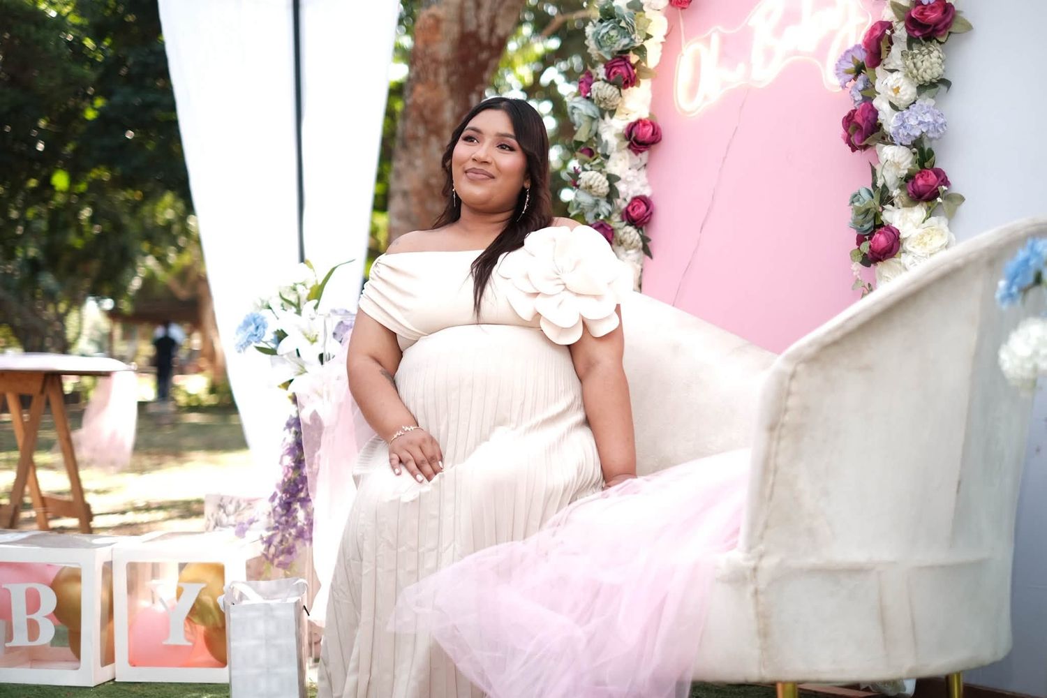Pregnant woman in white dress sitting on white chair at outdoor baby shower with floral decorations.