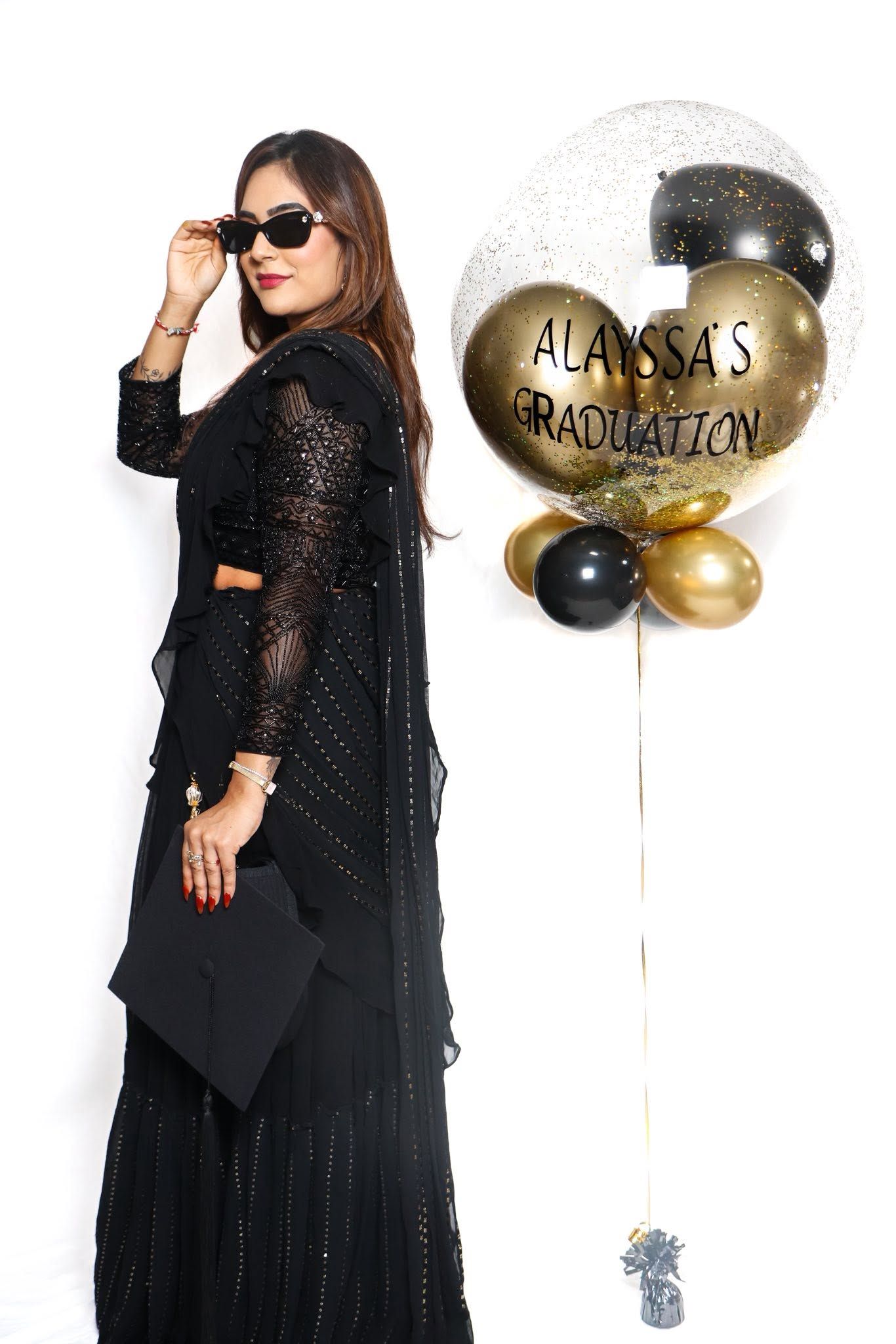 Woman in black sparkly outfit holding graduation cap poses with Alayssa's Graduation balloons in gold and black.