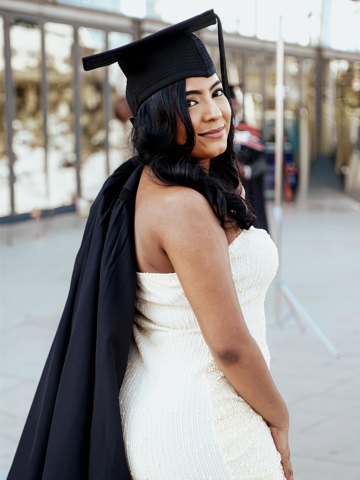 Woman in white strapless dress and black graduation cap and gown posing outdoors, looking over her shoulder.