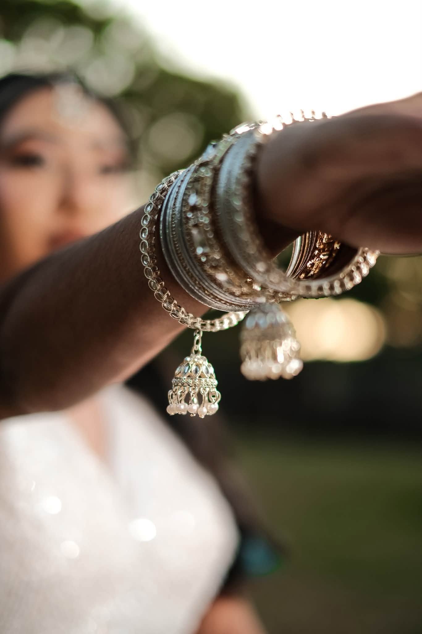 Close-up of a woman's wrist adorned with silver bangles and a jhumka charm, with a blurred bride in the background.