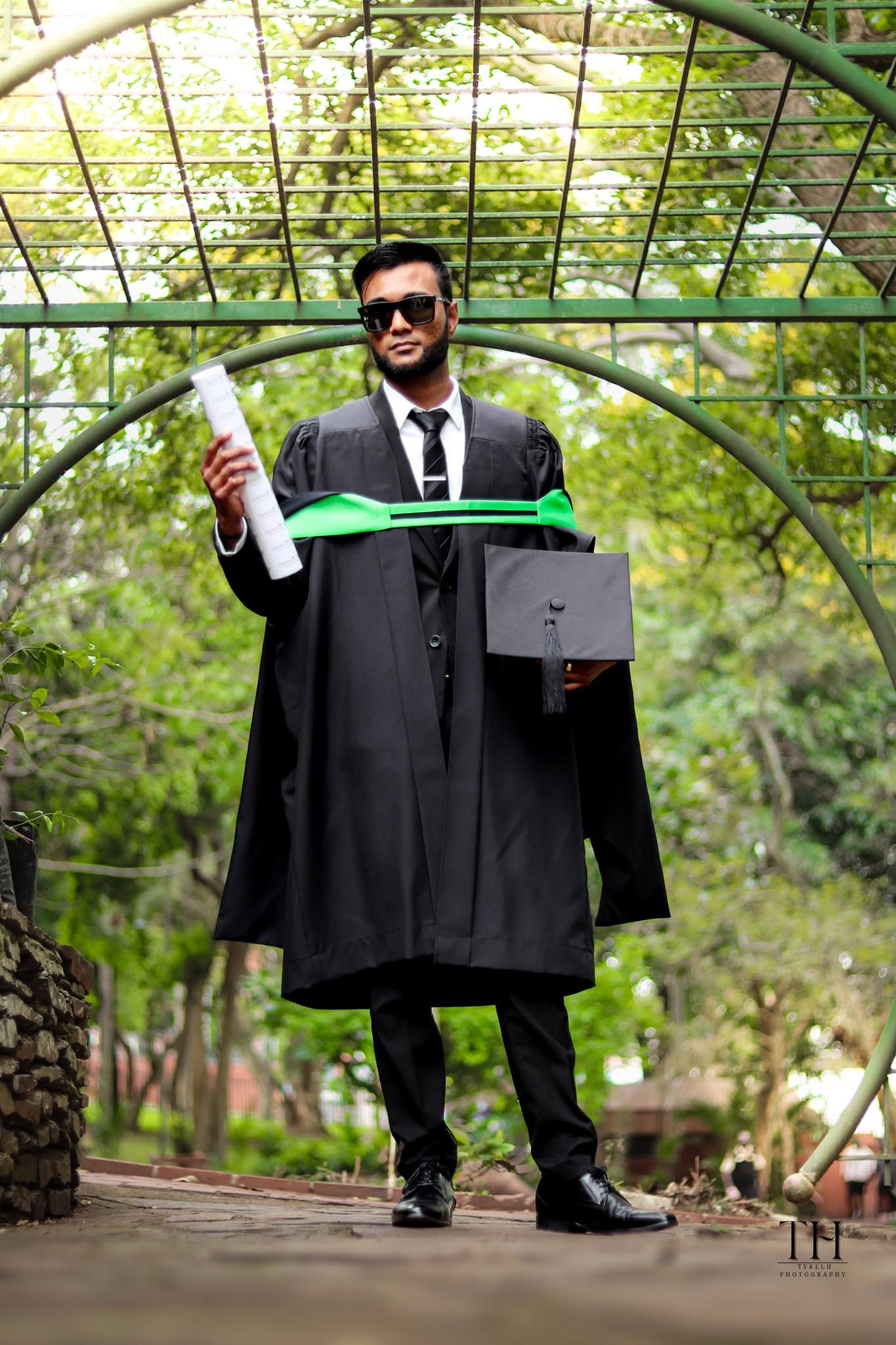 Graduate in black academic gown holding diploma and mortarboard cap, posing under green arch in park.