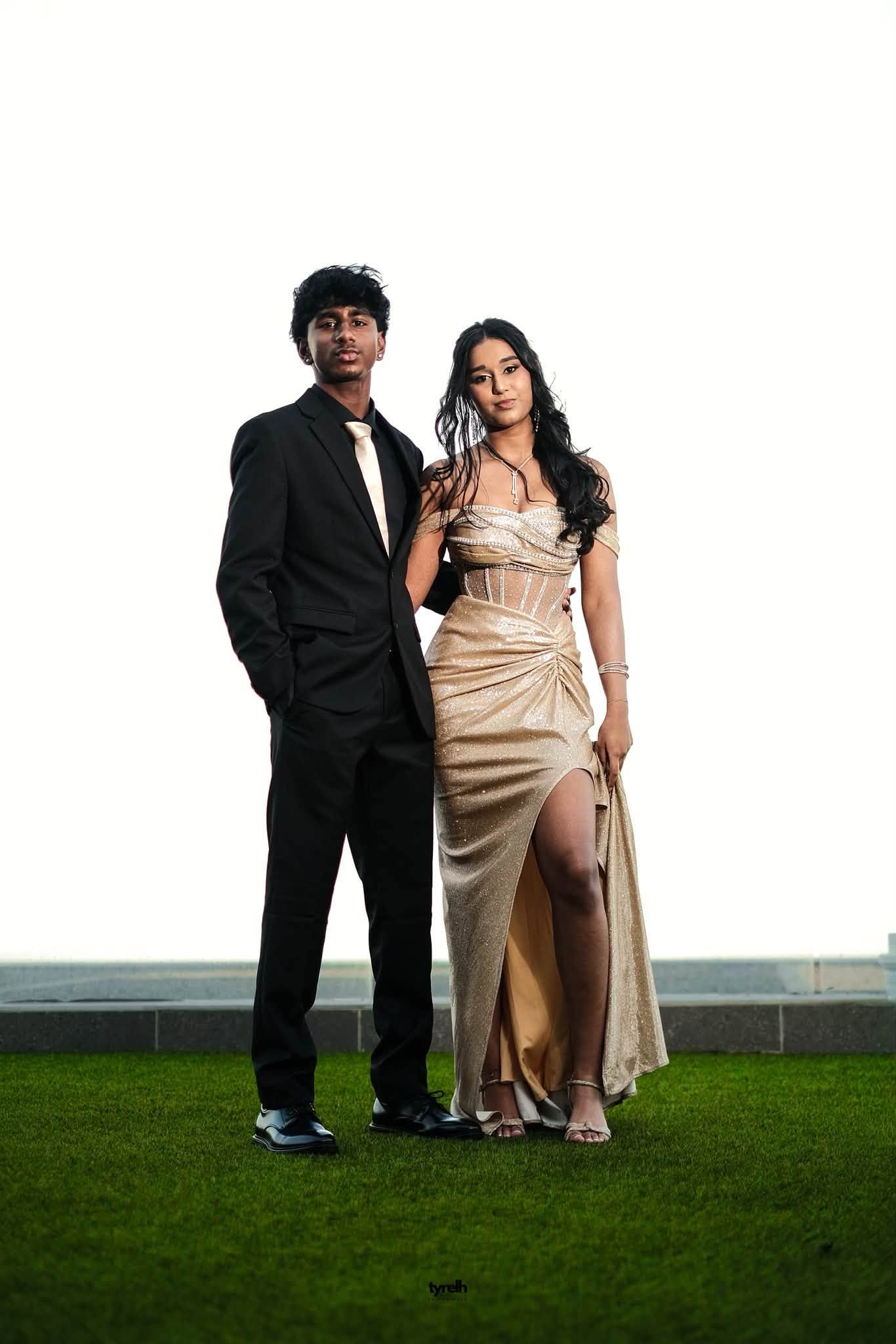 Elegant couple posing outdoors, man in black suit, woman in gold satin gown with corset detail on green lawn.
