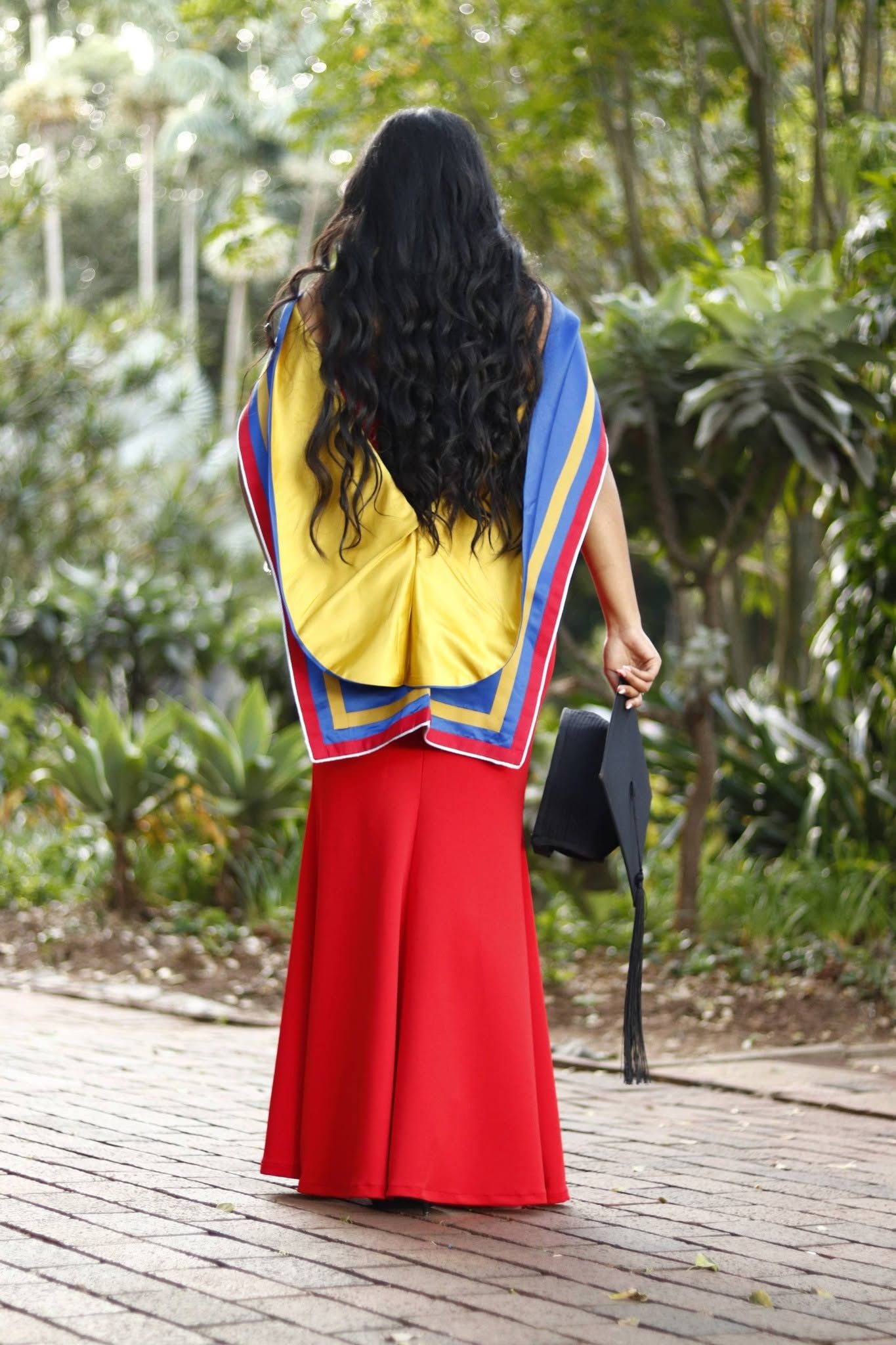 Woman in red skirt and yellow graduation gown holds mortarboard, walking through lush green garden path.