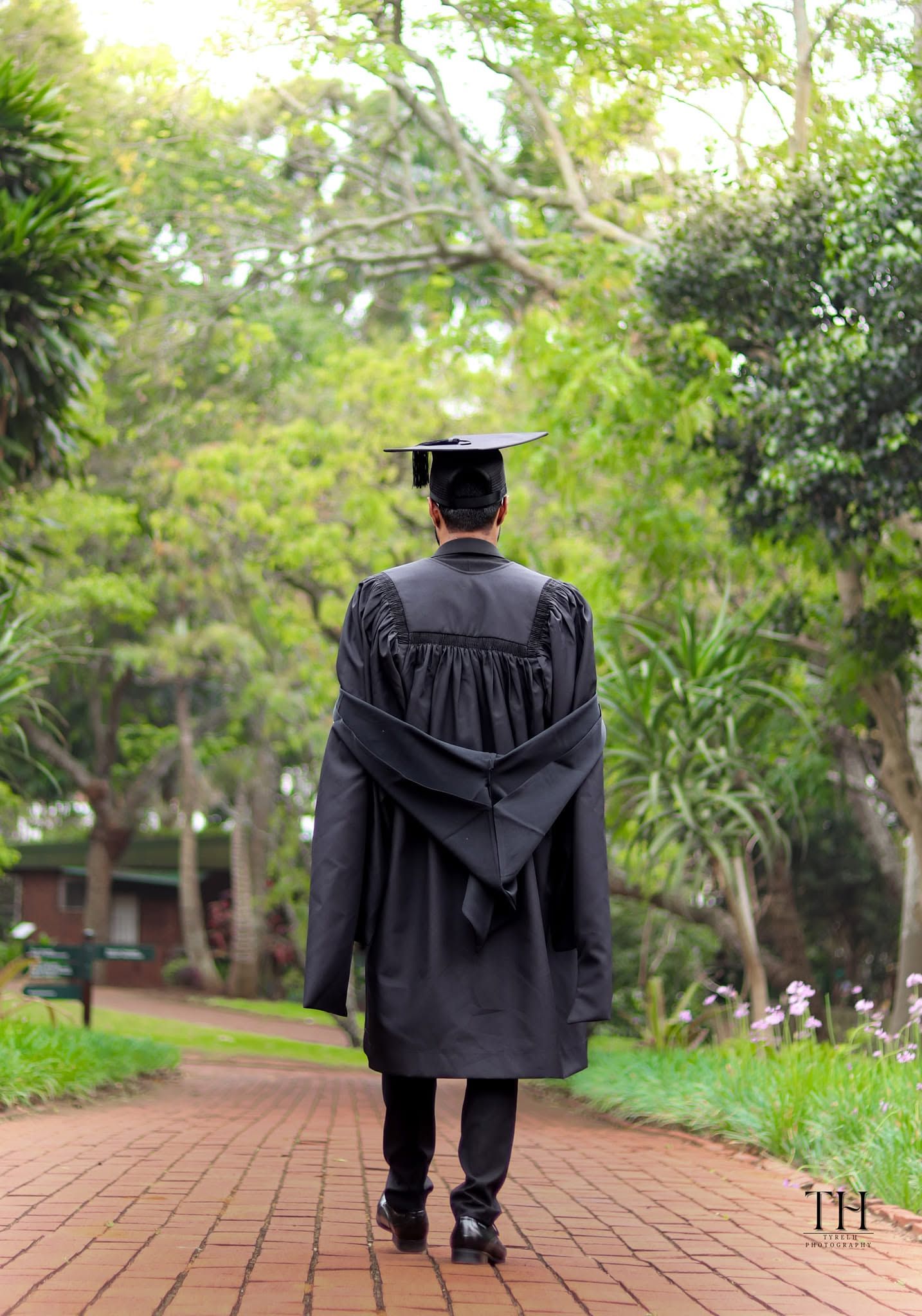Graduate in black cap and gown walks along a brick path through lush green trees.