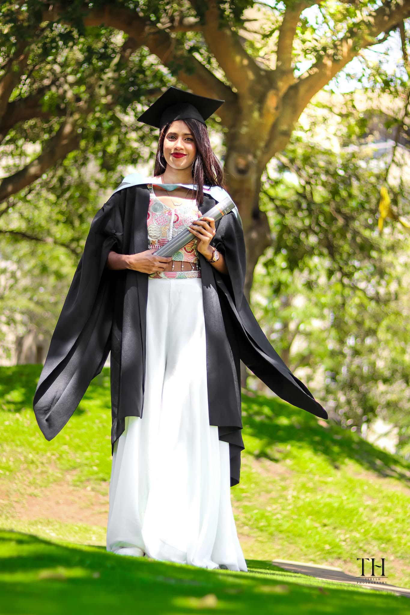 Young woman in black graduation gown and cap holding diploma outdoors on green campus grounds.