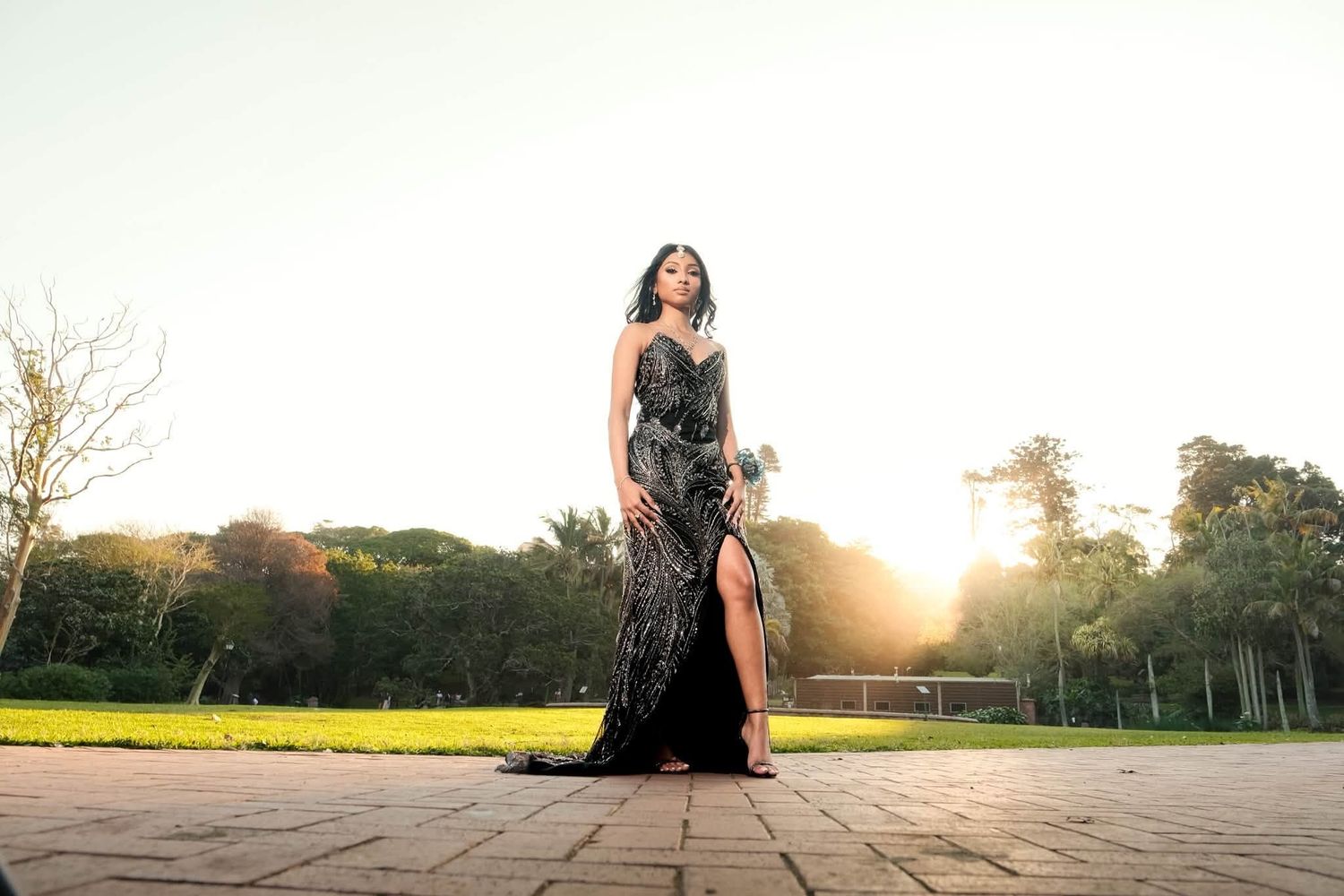 Woman in elegant black sequined gown poses outdoors at sunset with lush green trees in background.