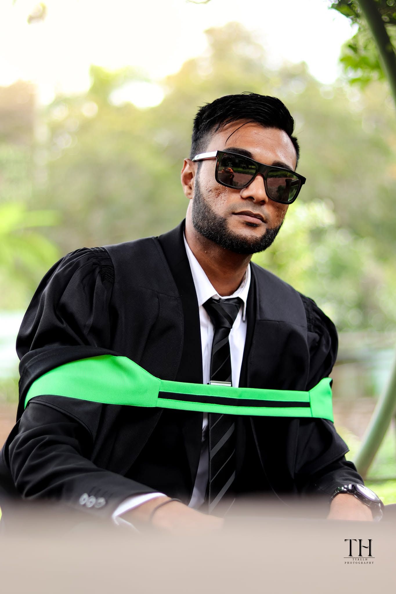 Graduate in black academic gown with green sash and sunglasses posing outdoors at graduation ceremony.