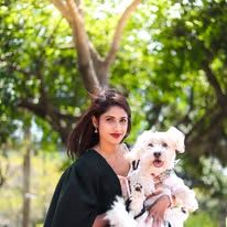 Woman in black outfit holding a fluffy white dog outdoors with lush green trees in the background.