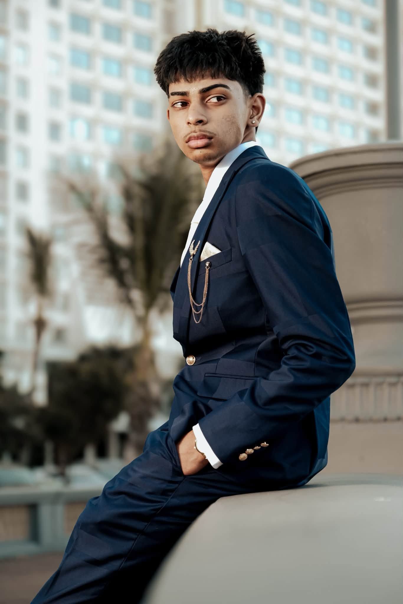 Young man in navy blue suit with gold chain brooch poses confidently outdoors near urban building.