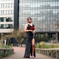 Woman in black sequin gown holding red roses on a wooden walkway outside a modern glass building.