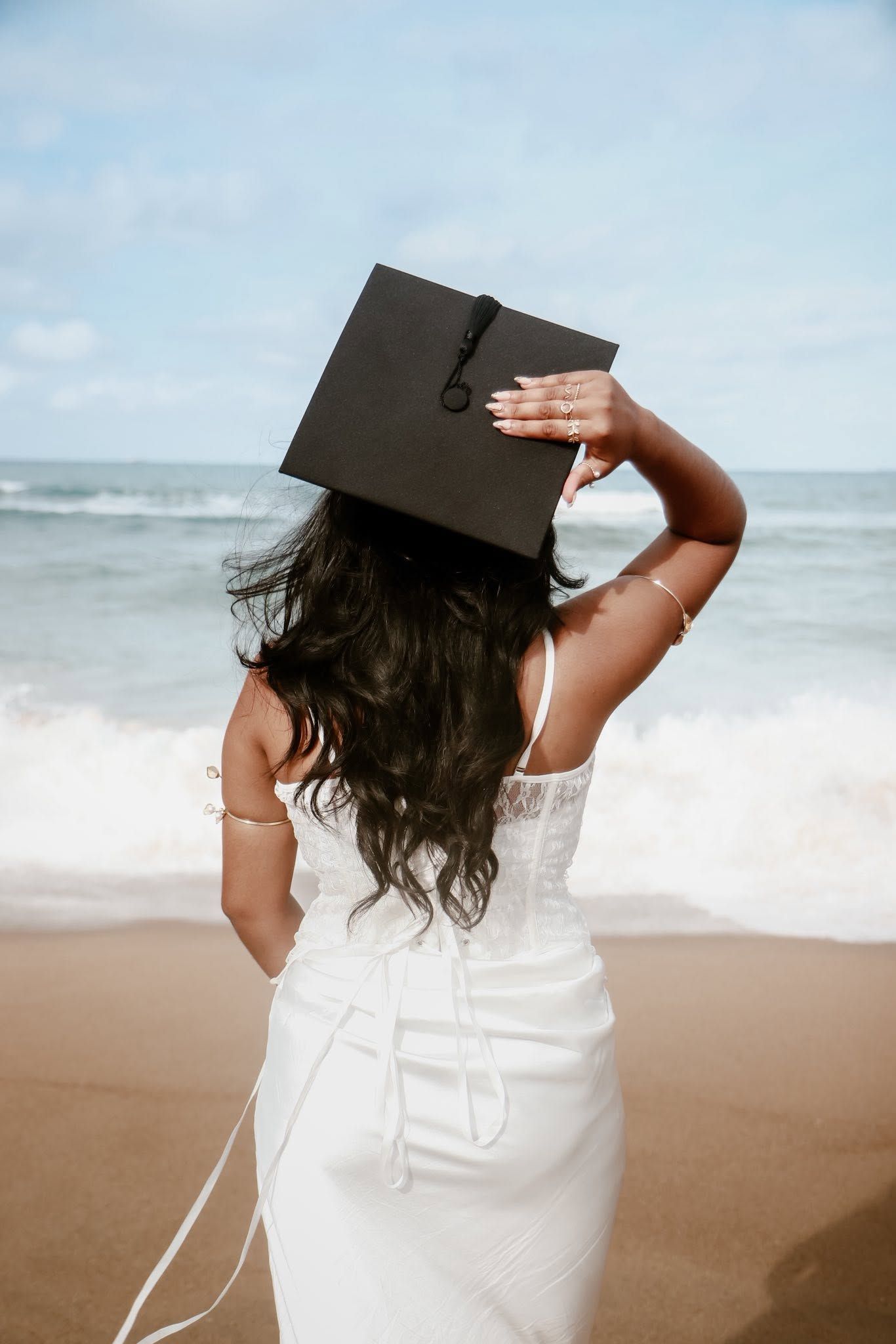 Woman in white dress holding graduation cap on a sunny beach with ocean waves in the background.