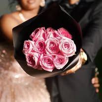 A bouquet of pink roses wrapped in black paper held by a person in formal attire.