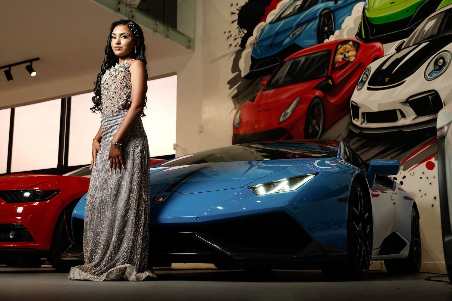 Woman in silver gown posing beside a blue Lamborghini Huracan in a luxury car showroom.