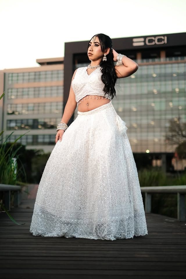 Young woman in elegant white embroidered lehenga choli with silver jewelry poses outdoors near modern building.