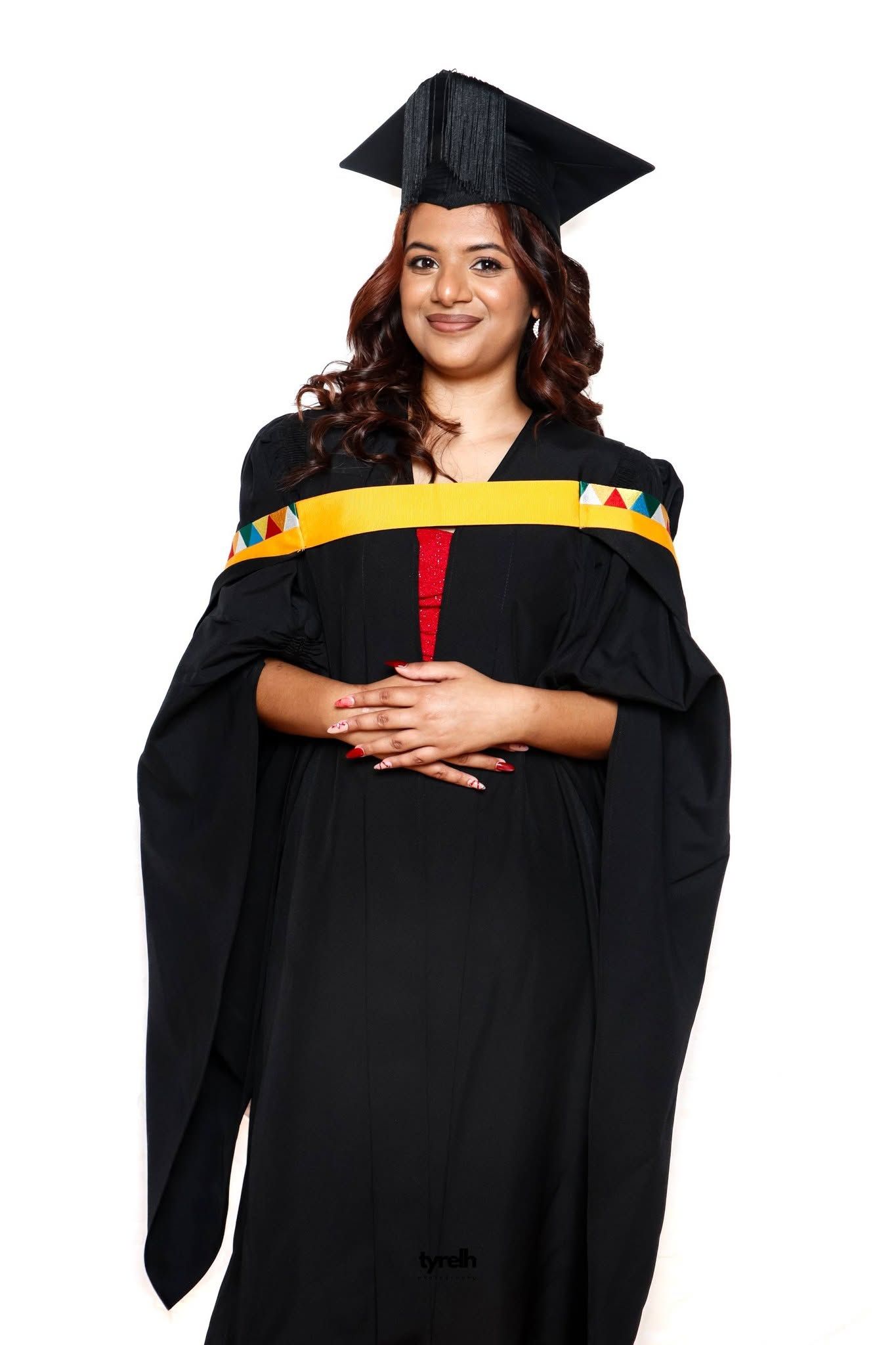 Woman in black graduation gown and mortarboard cap with colorful sash smiling against white background.
