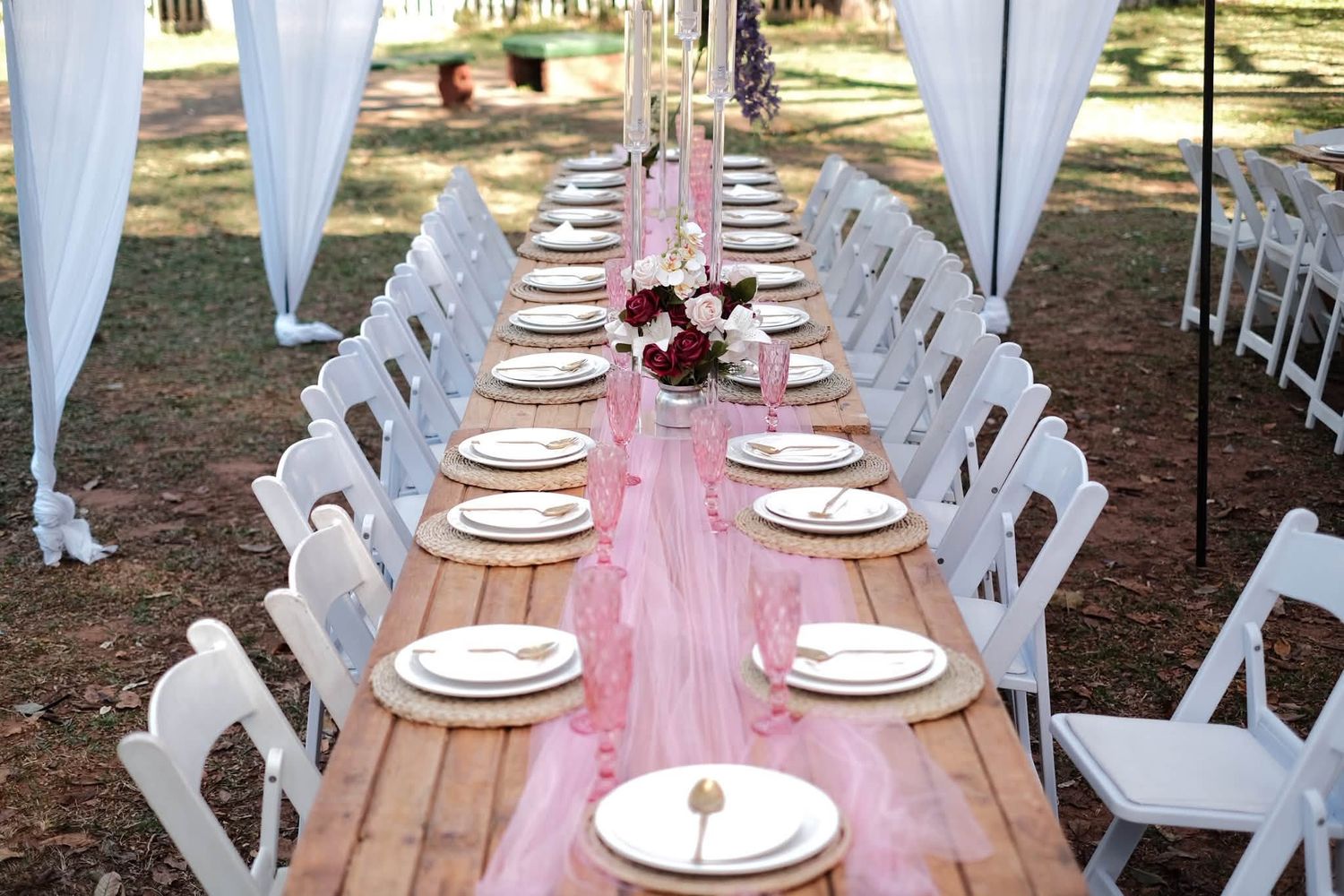 Elegant outdoor wedding reception table with pink runner, white chairs, gold place settings, and floral centerpieces.