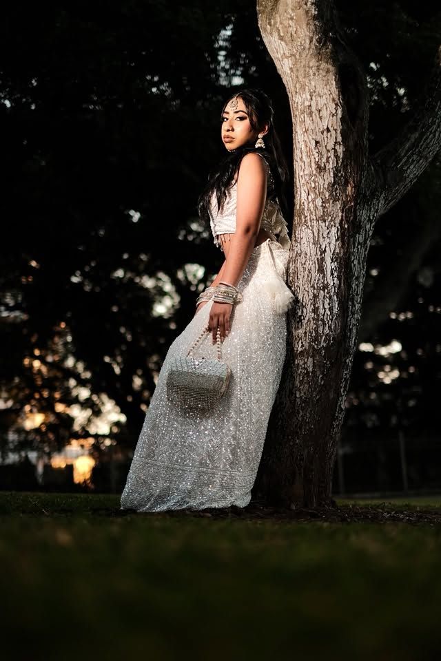 Young woman in white sequin gown leans against tree at dusk, holding sparkly clutch in outdoor park setting.