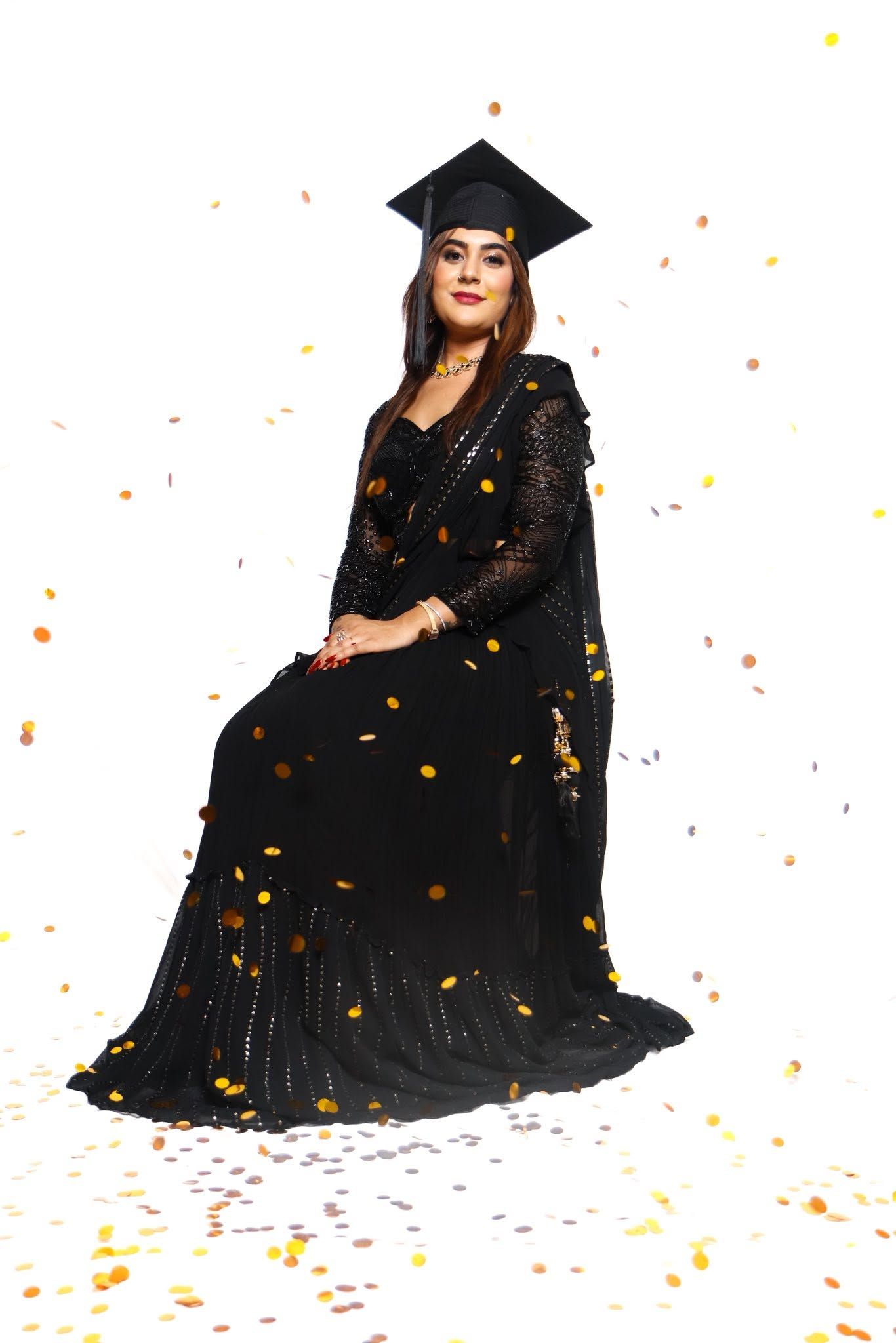 Woman in black saree and graduation cap sits elegantly amid falling gold confetti on white background.