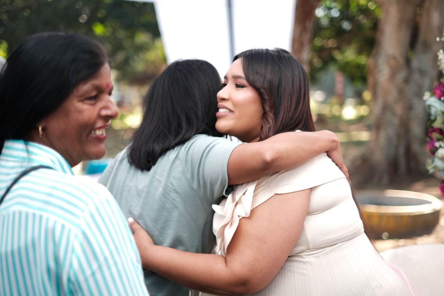 Three women sharing warm hugs outdoors, smiling joyfully in a sunny garden setting.