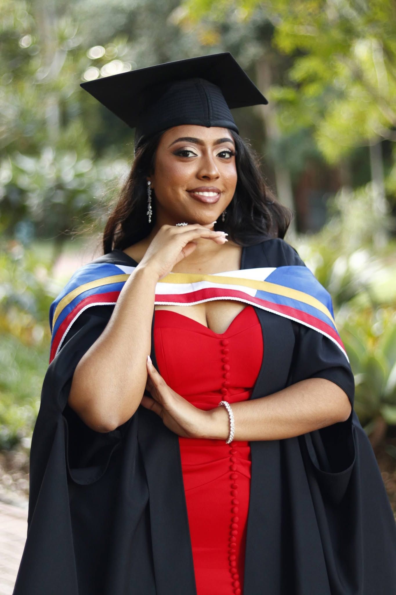 Smiling graduate woman wearing black cap and gown with colorful hood and red dress posing outdoors.