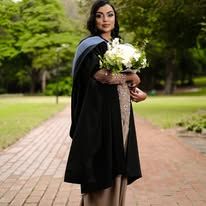 Woman in black graduation gown holding white bouquet on brick path surrounded by green trees.